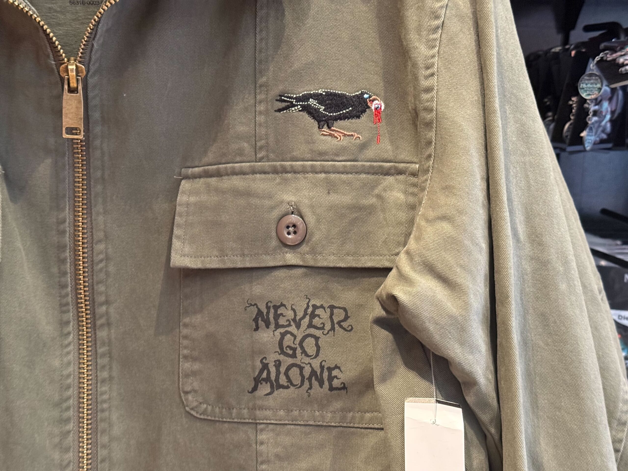Close-up of a khaki jacket pocket from HHN Merch 2025, with "NEVER GO ALONE" and an embroidered bird with blood on its beak.
