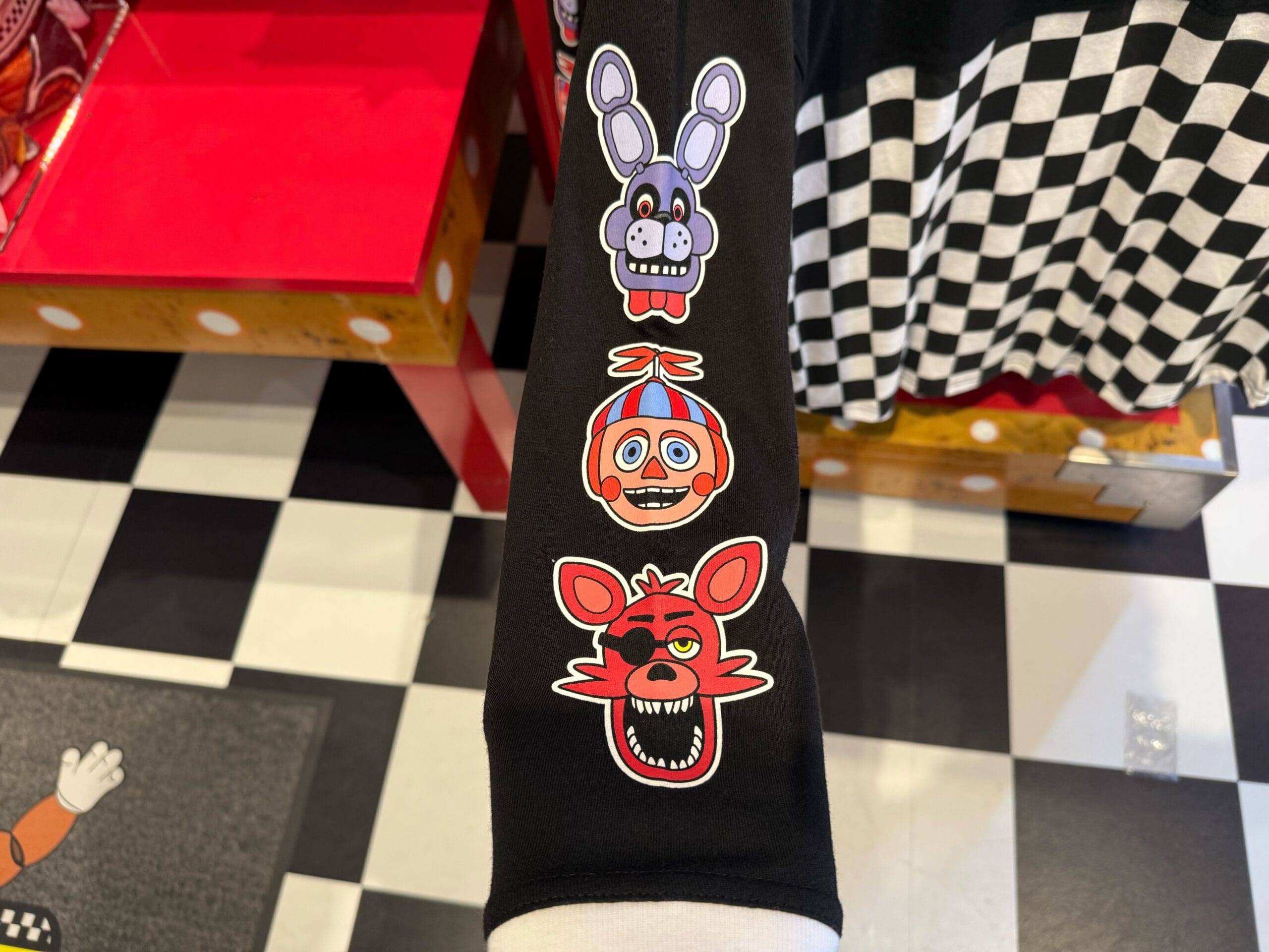 Three cartoon animatronic faces on a black sleeve, inspired by HHN Merch 2025, over a checkered floor background.