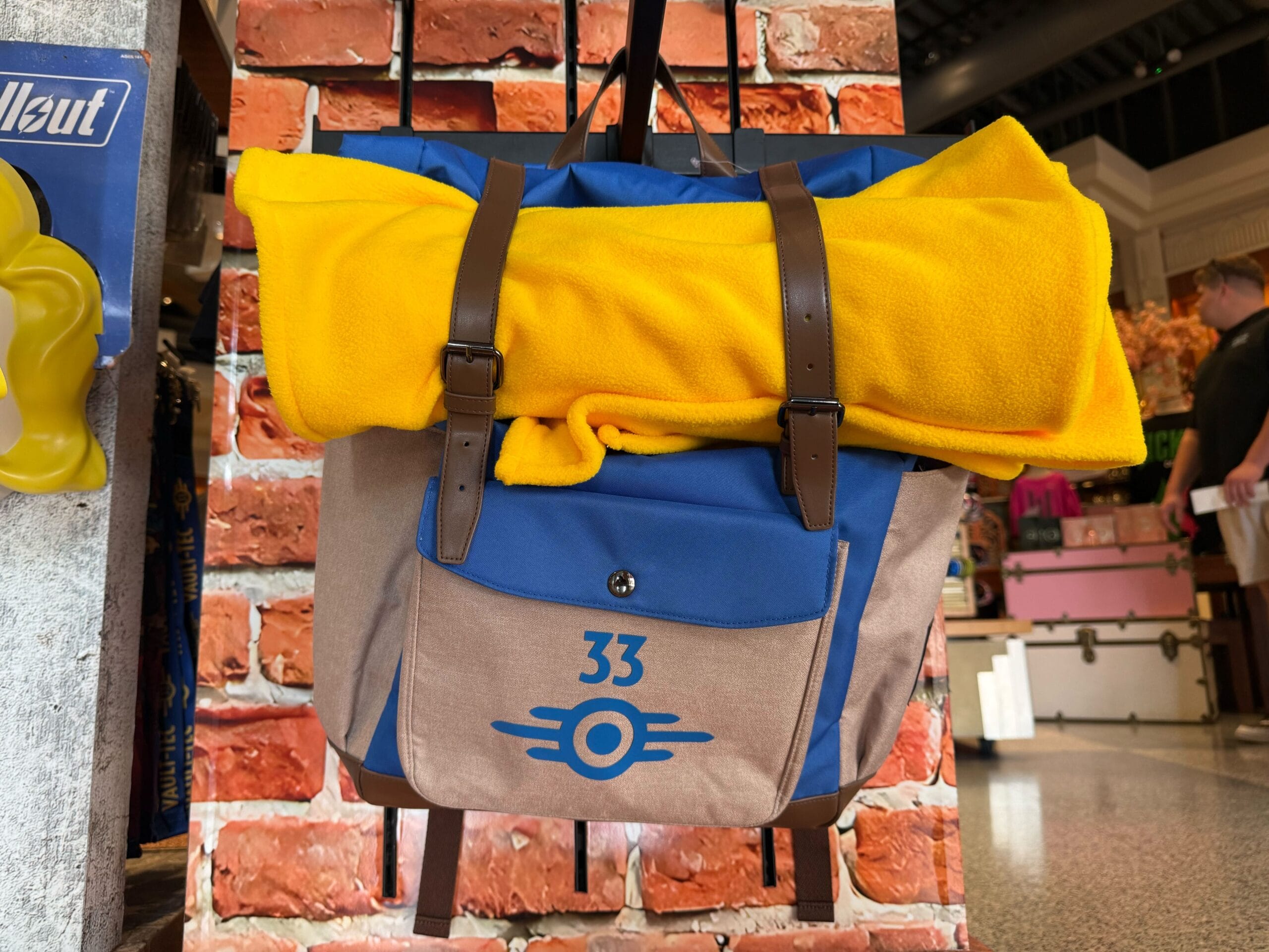 A blue and tan backpack with 33 and a yellow blanket, HHN Merch 2025, hangs in front of a brick wall.