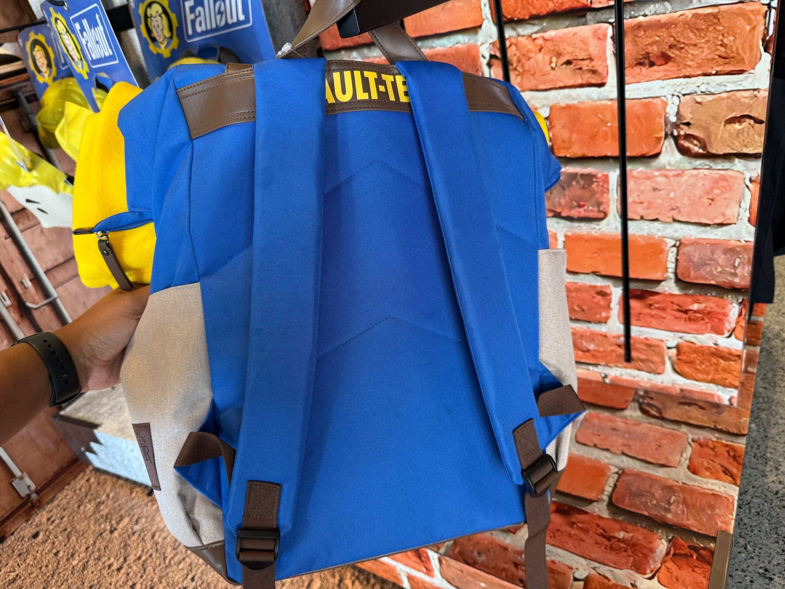 A blue and yellow "Vault-Tec" backpack from Fallout, shown at Universal Studios Hollywood in front of a brick wall display.