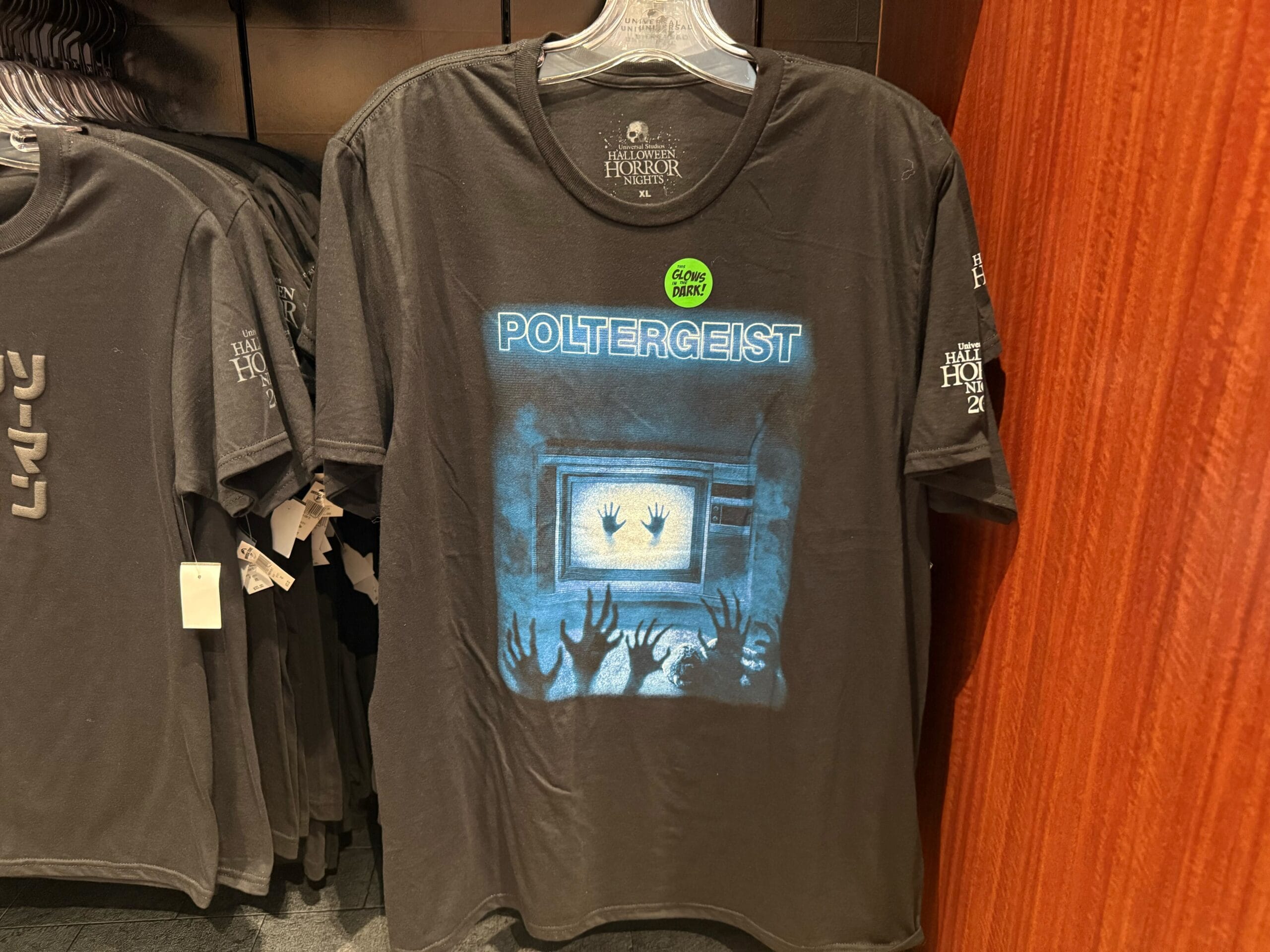 Black "Poltergeist" T-shirt, part of HHN Merch 2025, with hands reaching for a glowing TV, displayed in-store.