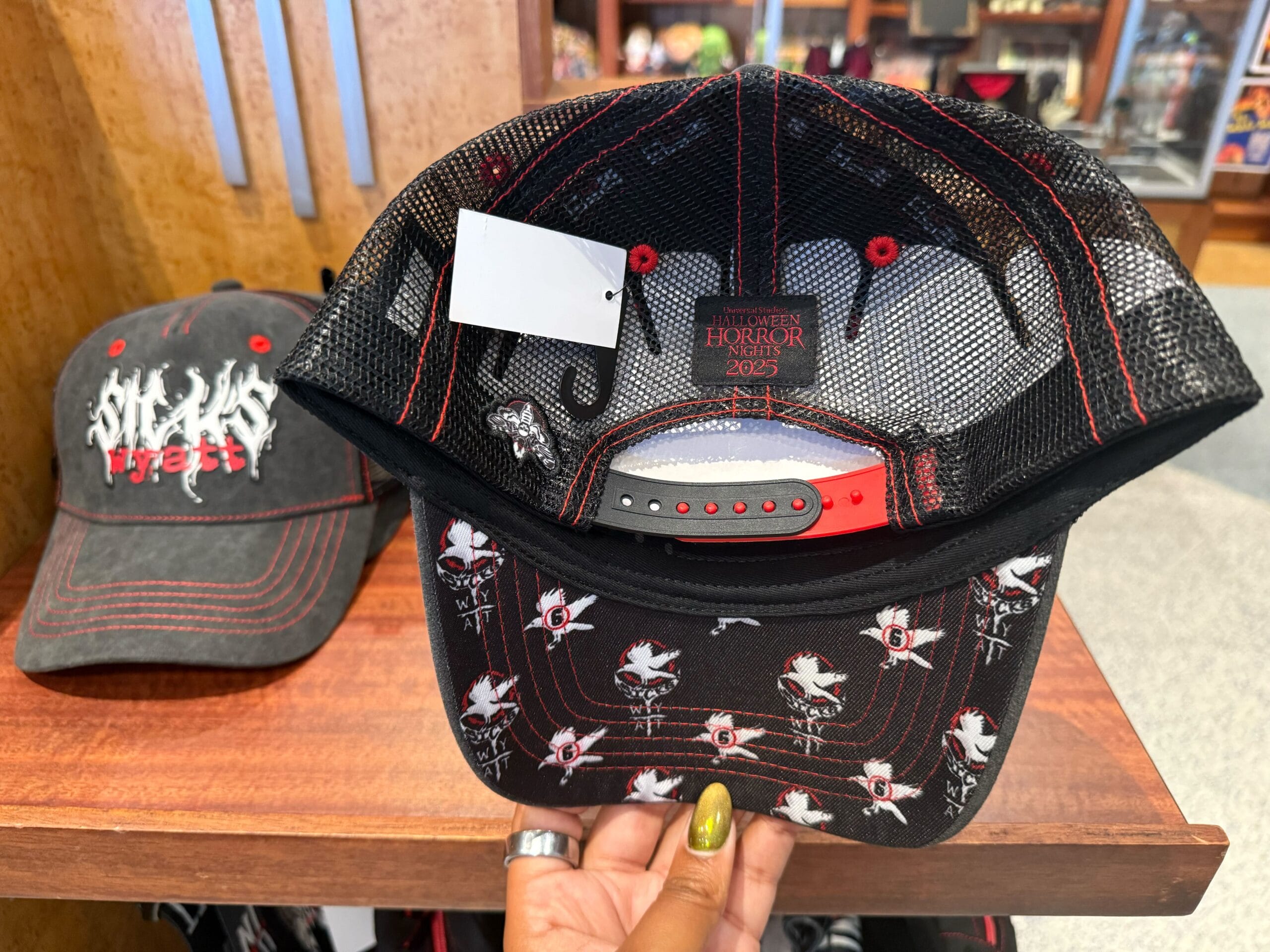 A hand holds a black cap with white doves and "6"; another cap nearby features Halloween Horror Nights Merchandise.