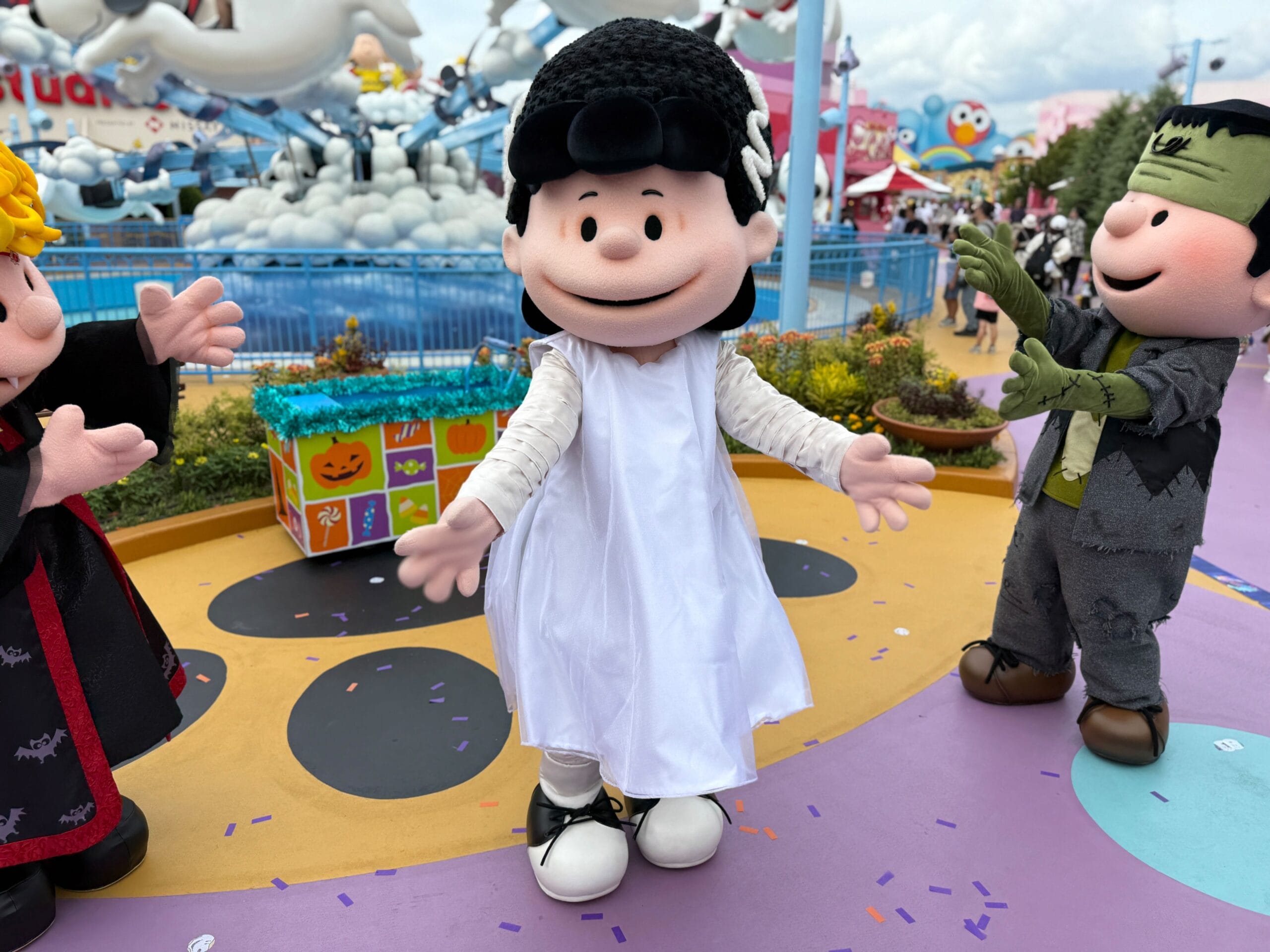 A person in a Lucy Van Pelt costume wears a white dress, posing outdoors with other Peanuts characters in Halloween outfits.