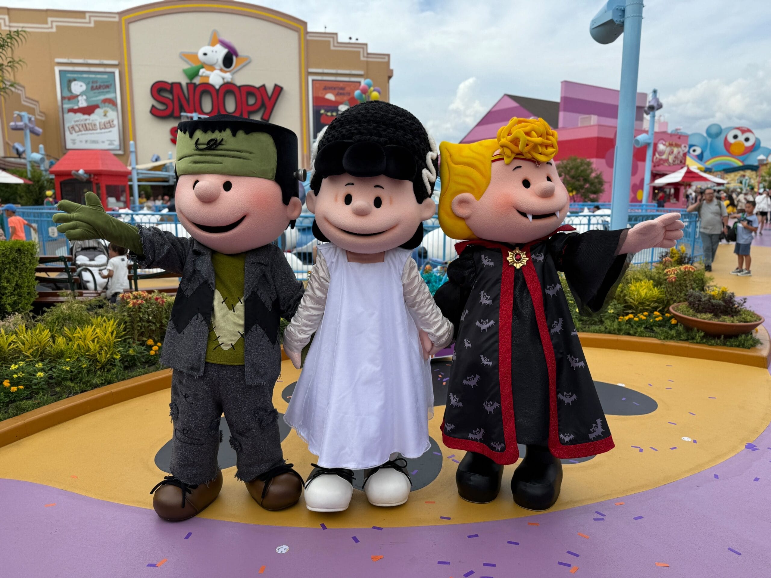 Three Peanuts characters in Halloween costumes stand outside a Snoopy-themed attraction at an amusement park.