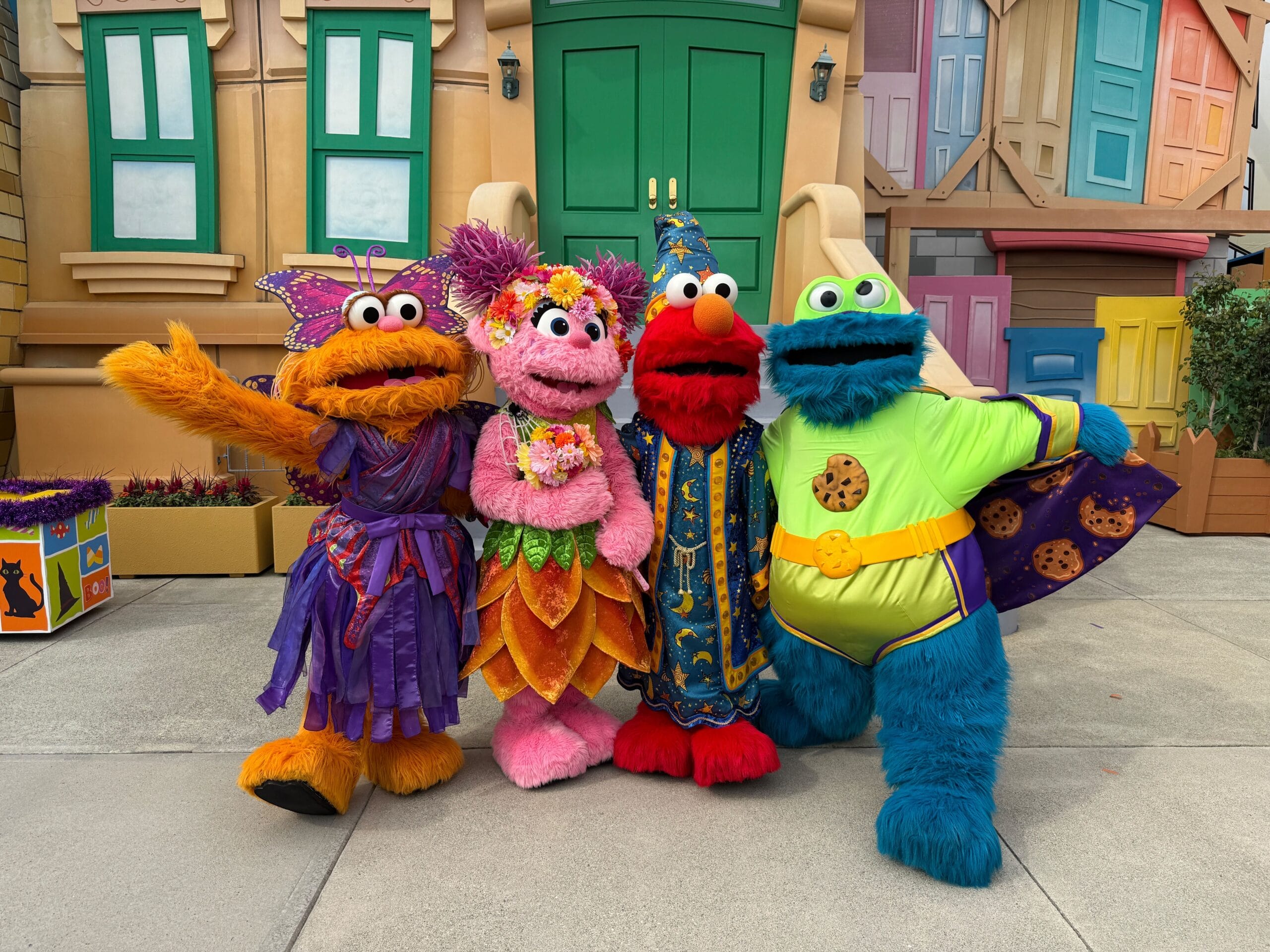 Four Sesame Street characters in colorful costumes pose together in front of a bright, cartoon-style building.