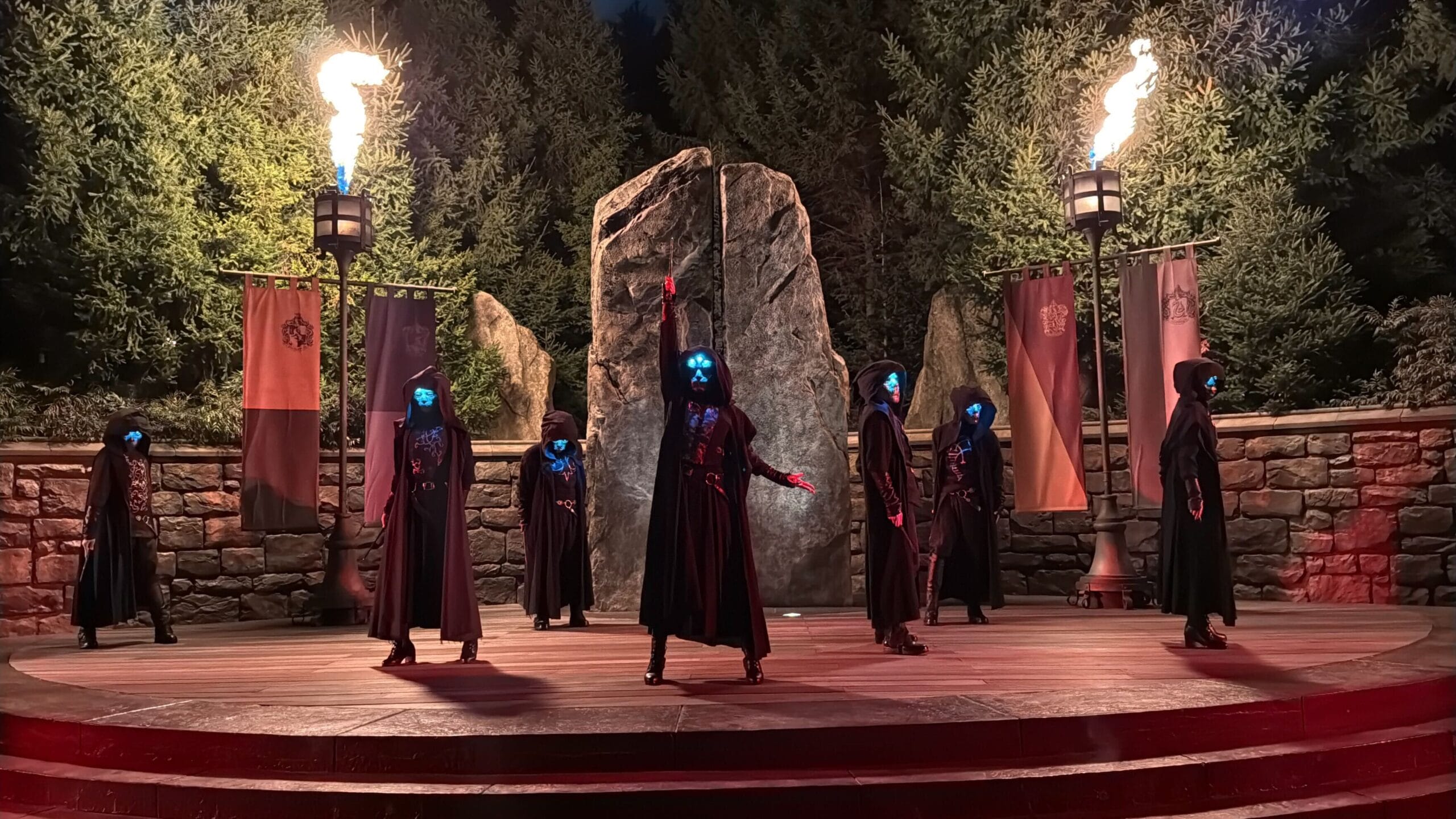 Seven hooded figures with glowing blue masks stand on a stage with fire torches and banners by a large stone at night.