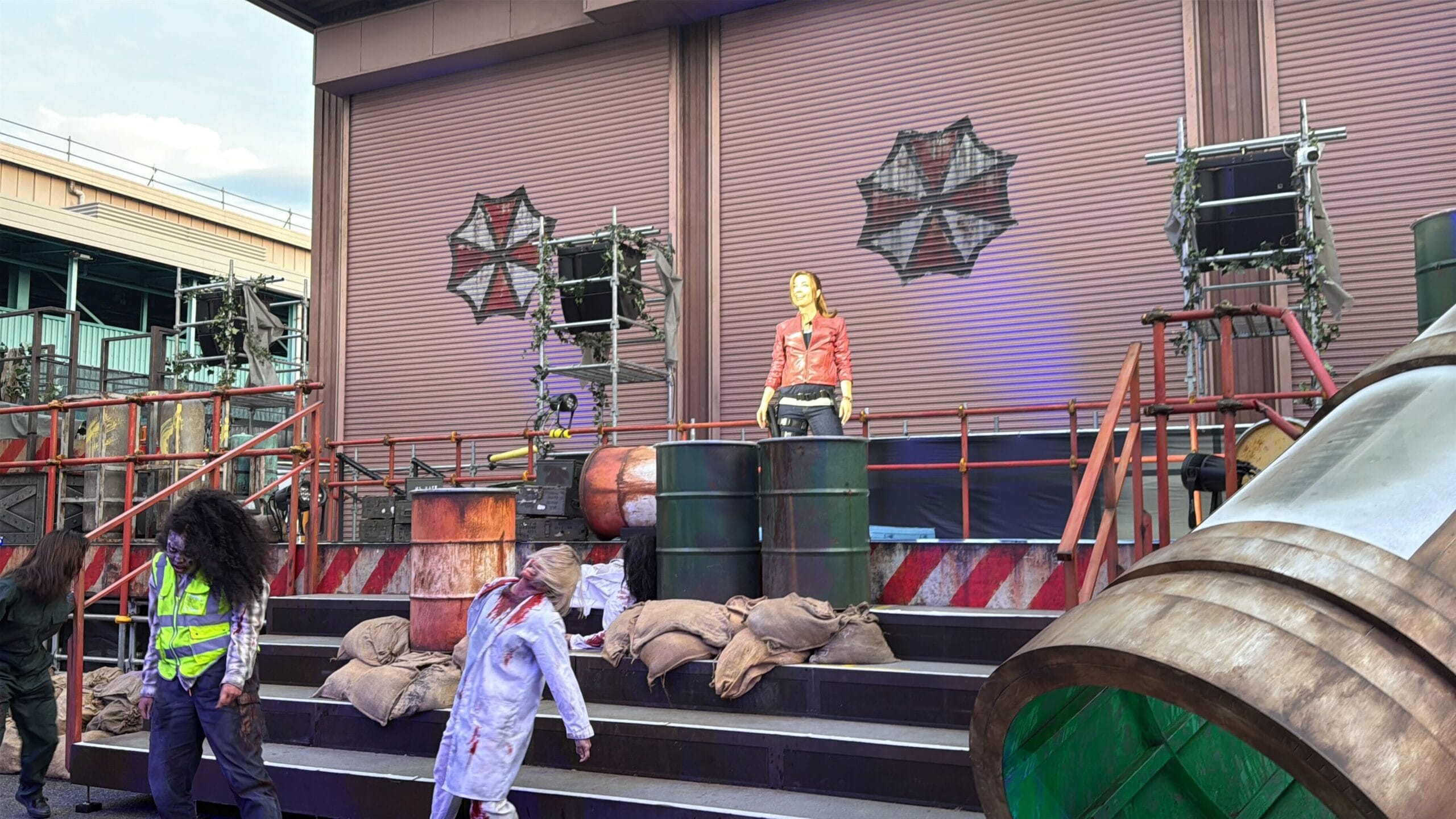 A person in a red jacket stands on a stage with barrels and sandbags; two people in costume walk in front.