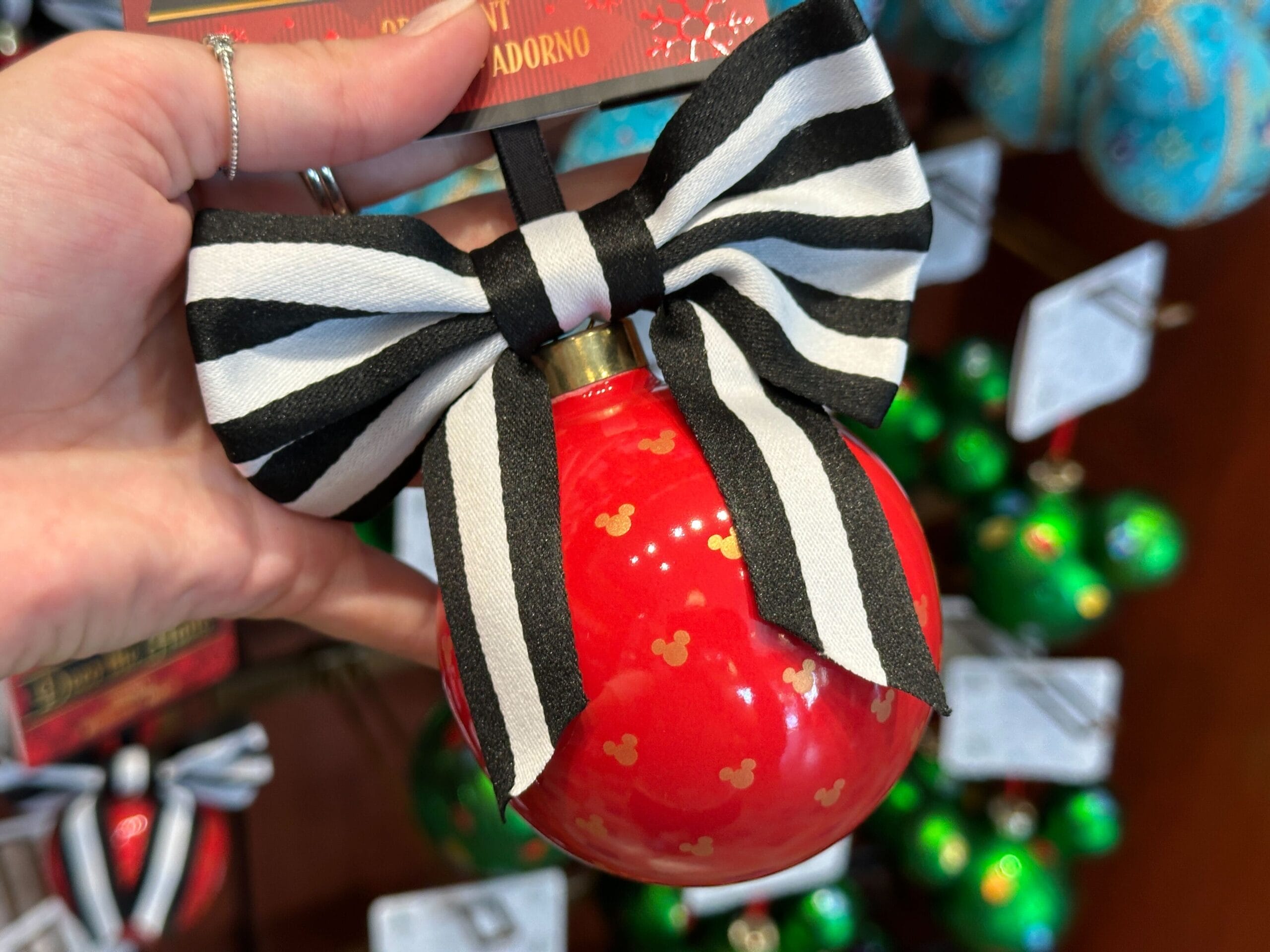 A hand holds a red Mickey Mouse holiday ornament with gold accents and a black-and-white striped bow.