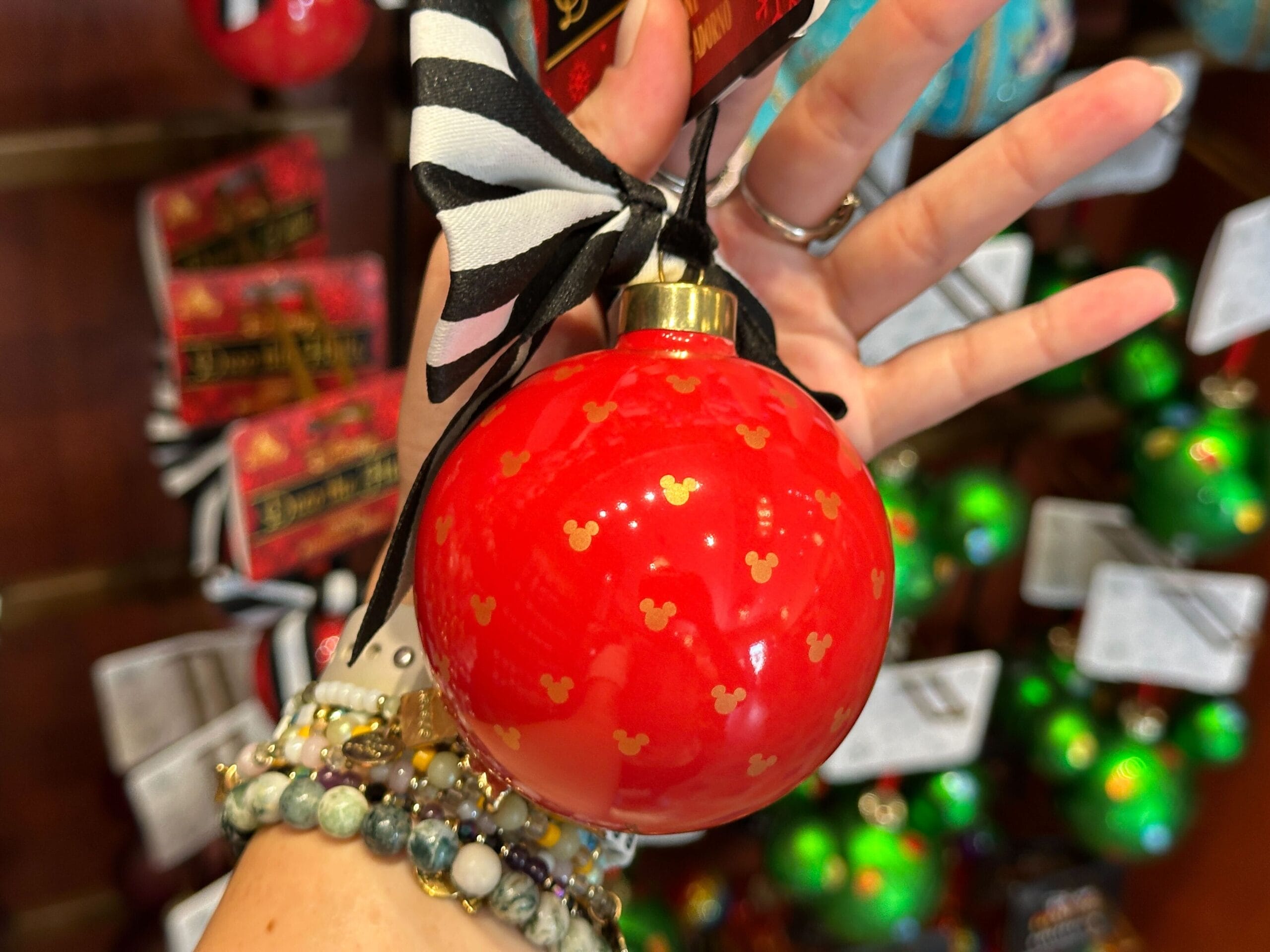 A hand holds a red Mickey Mouse holiday ornament with gold patterns and a black-and-white striped ribbon.