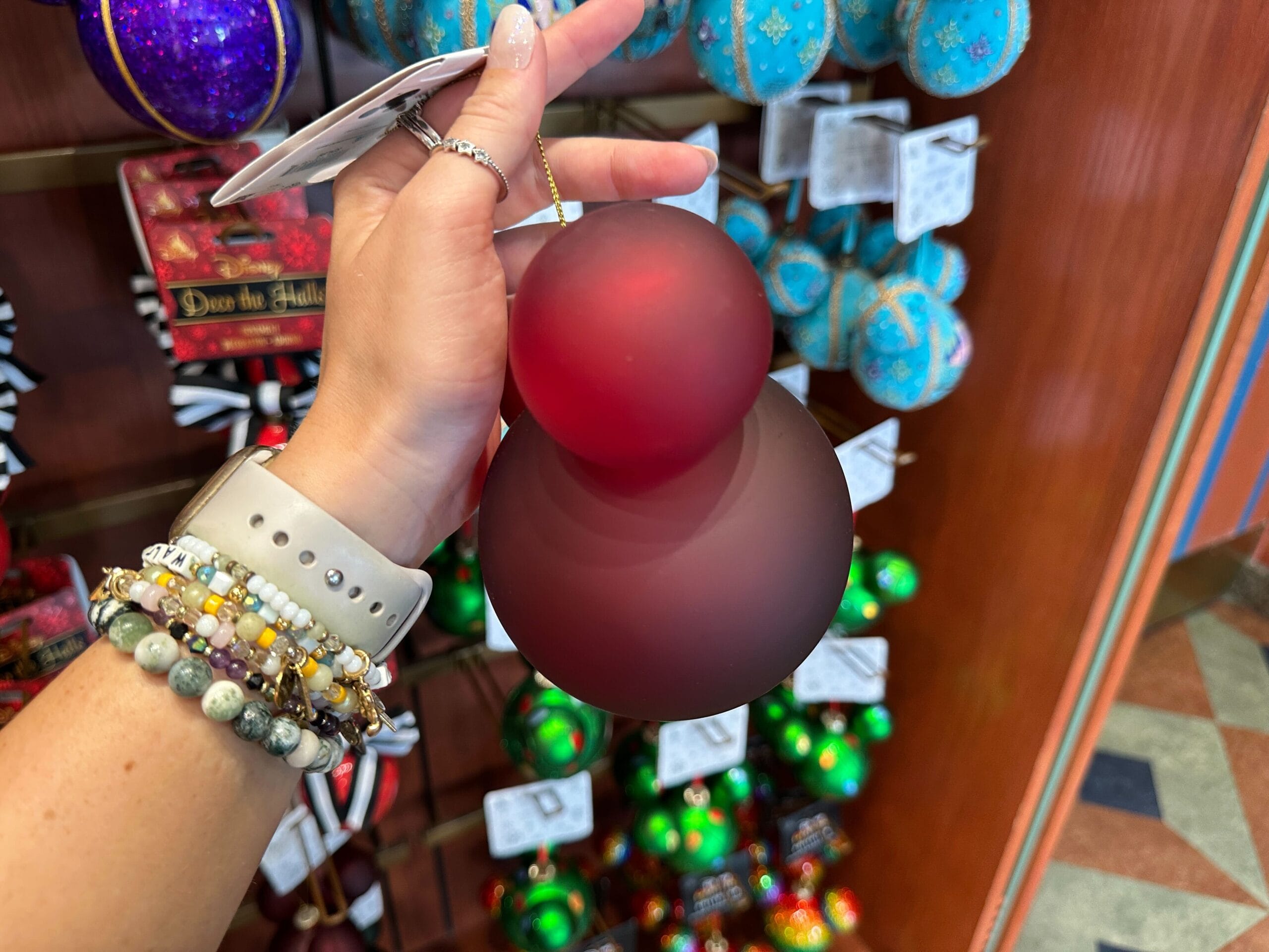 A hand holds a red, frosted Mickey Mouse-shaped ornament in a store filled with holiday decorations.