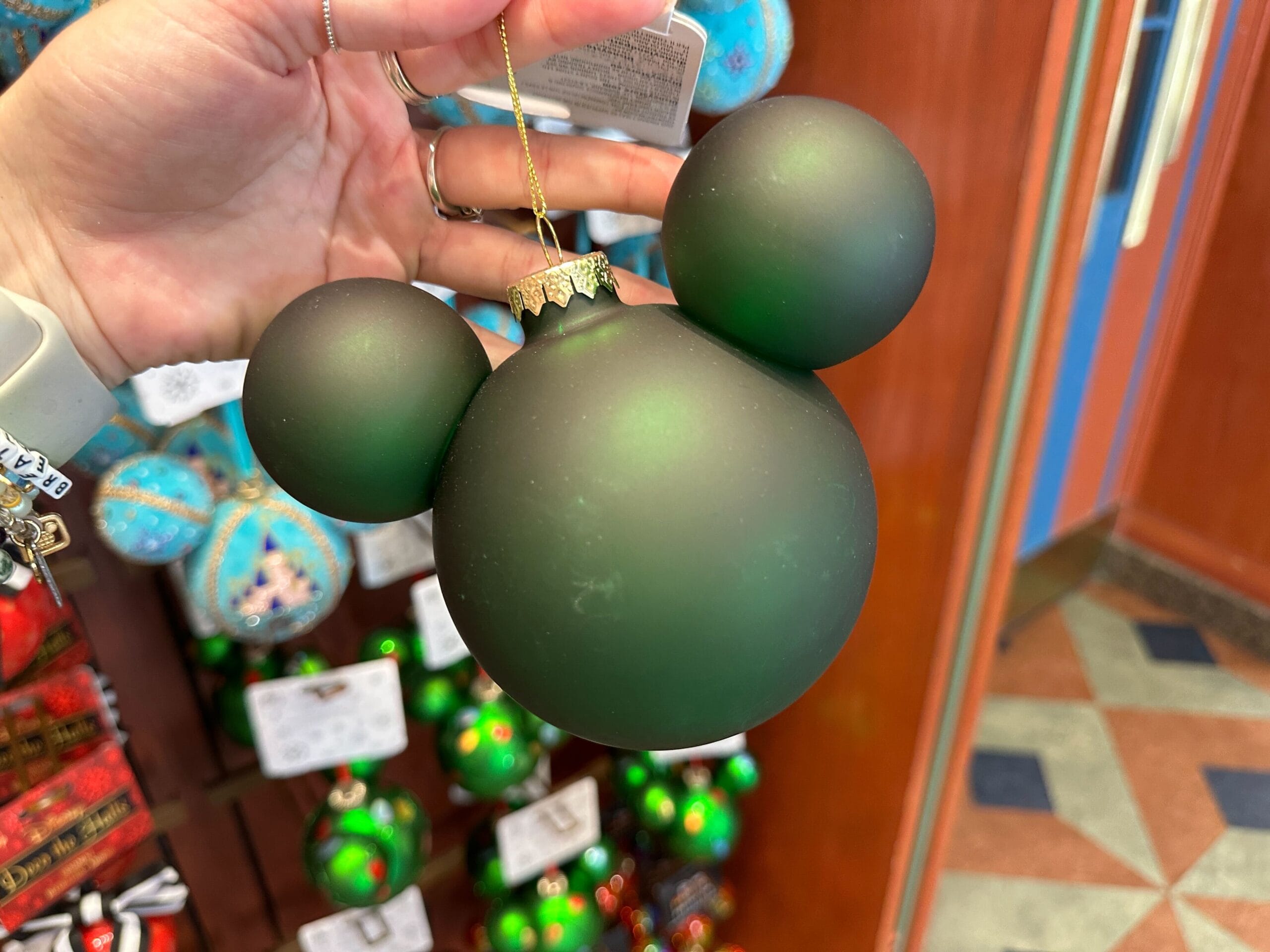 A hand holds a green Mickey Mouse-shaped holiday ornament in a store filled with festive Disney World decorations.