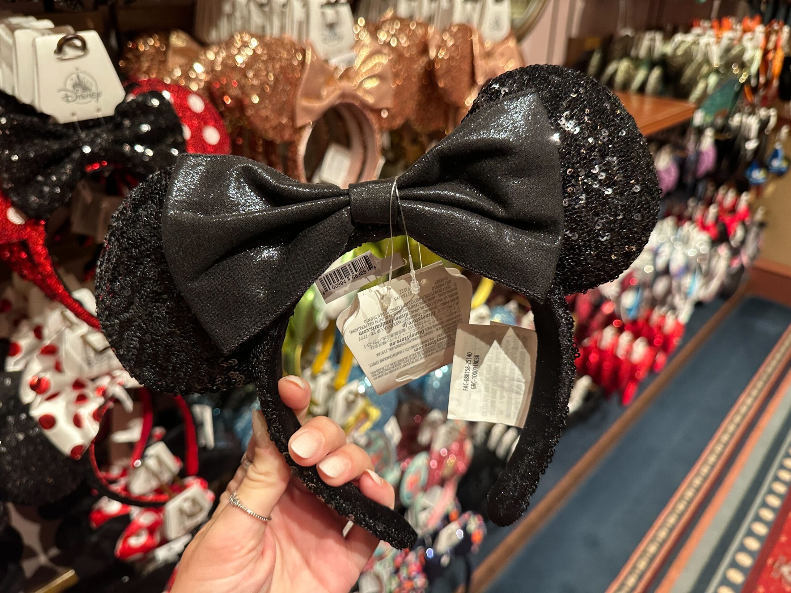 A hand holds a black sequined Minnie Mouse ear headband with a bow in a store display of various Disney-themed headbands.