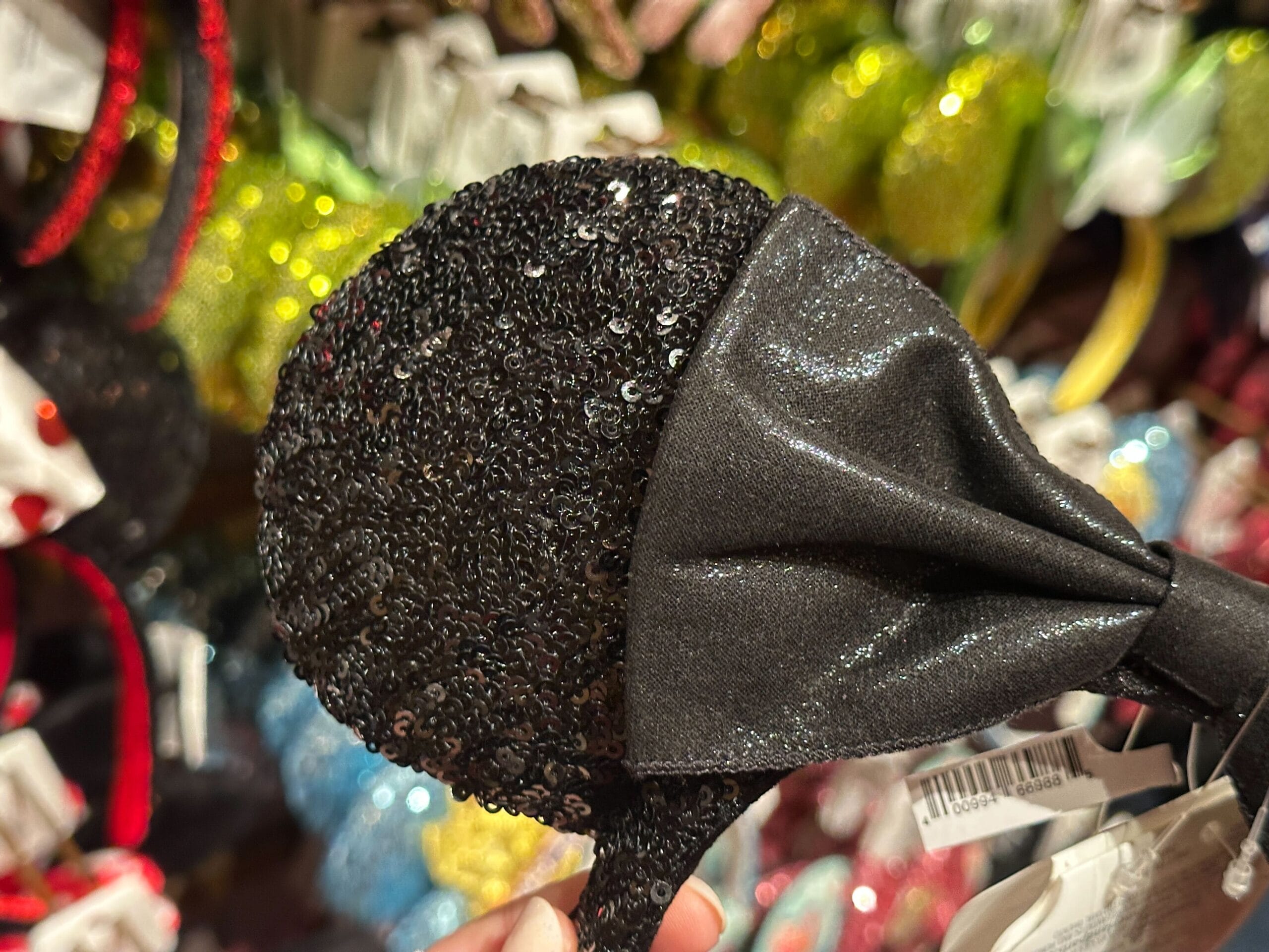 A hand holds a sparkly black sequin Mickey Mouse ear headband with a shiny black bow in a store display.