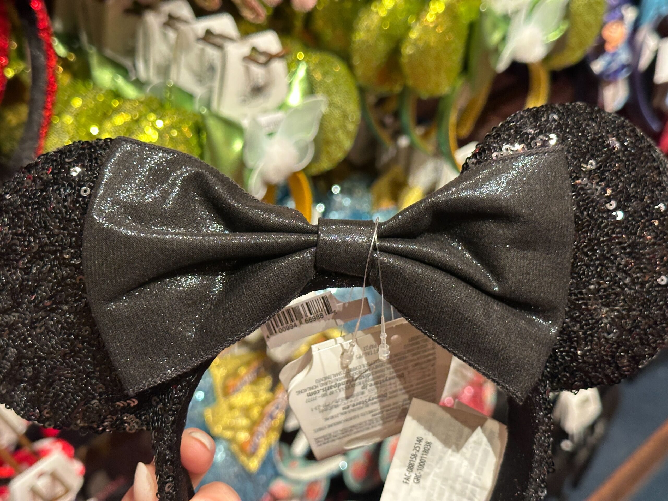 A sequined black headband with large black bow, displayed in a store with colorful headbands in the background.