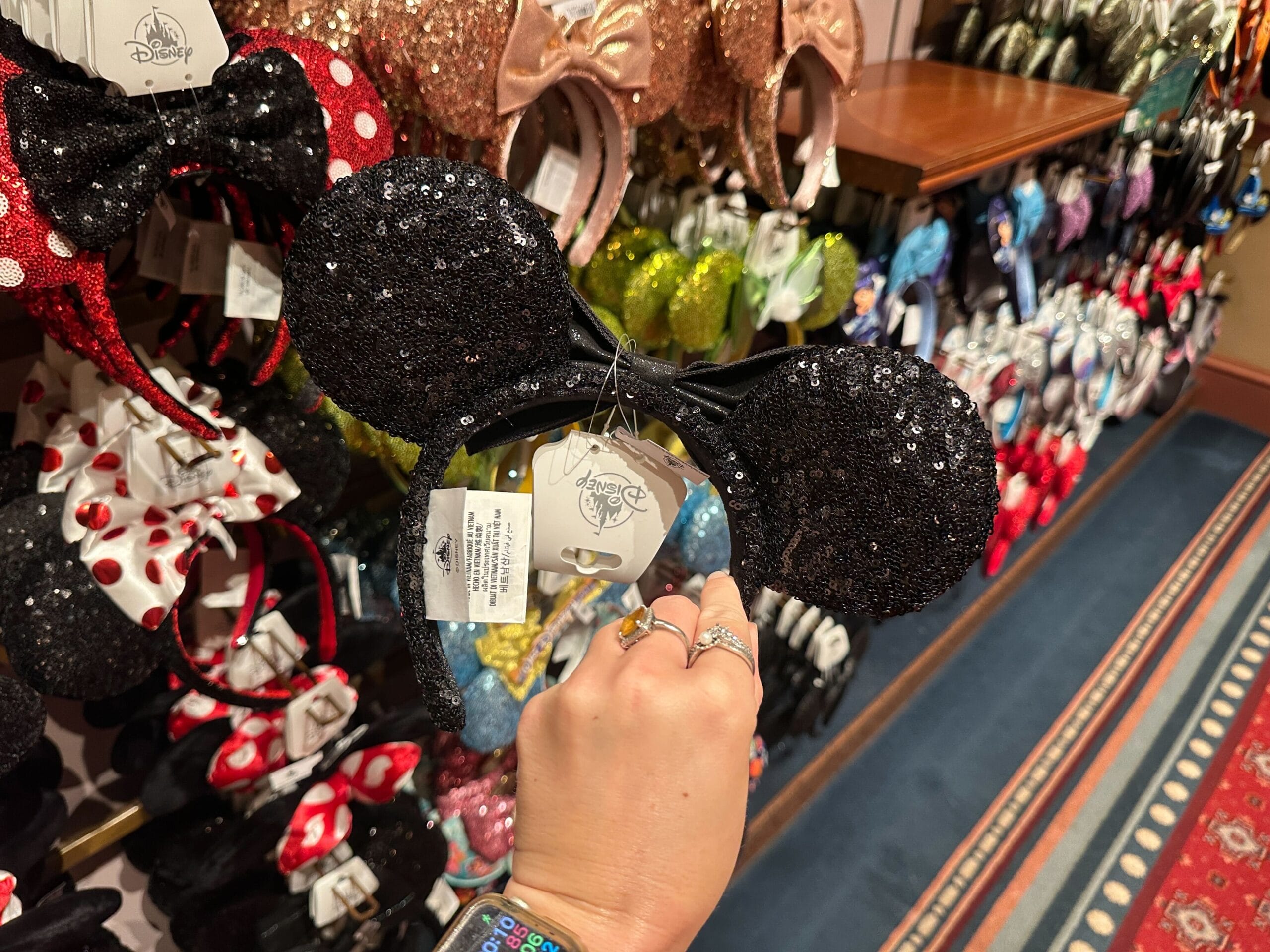 A hand holds black sequin Minnie Mouse ears in a store with various colorful headbands displayed on the wall.