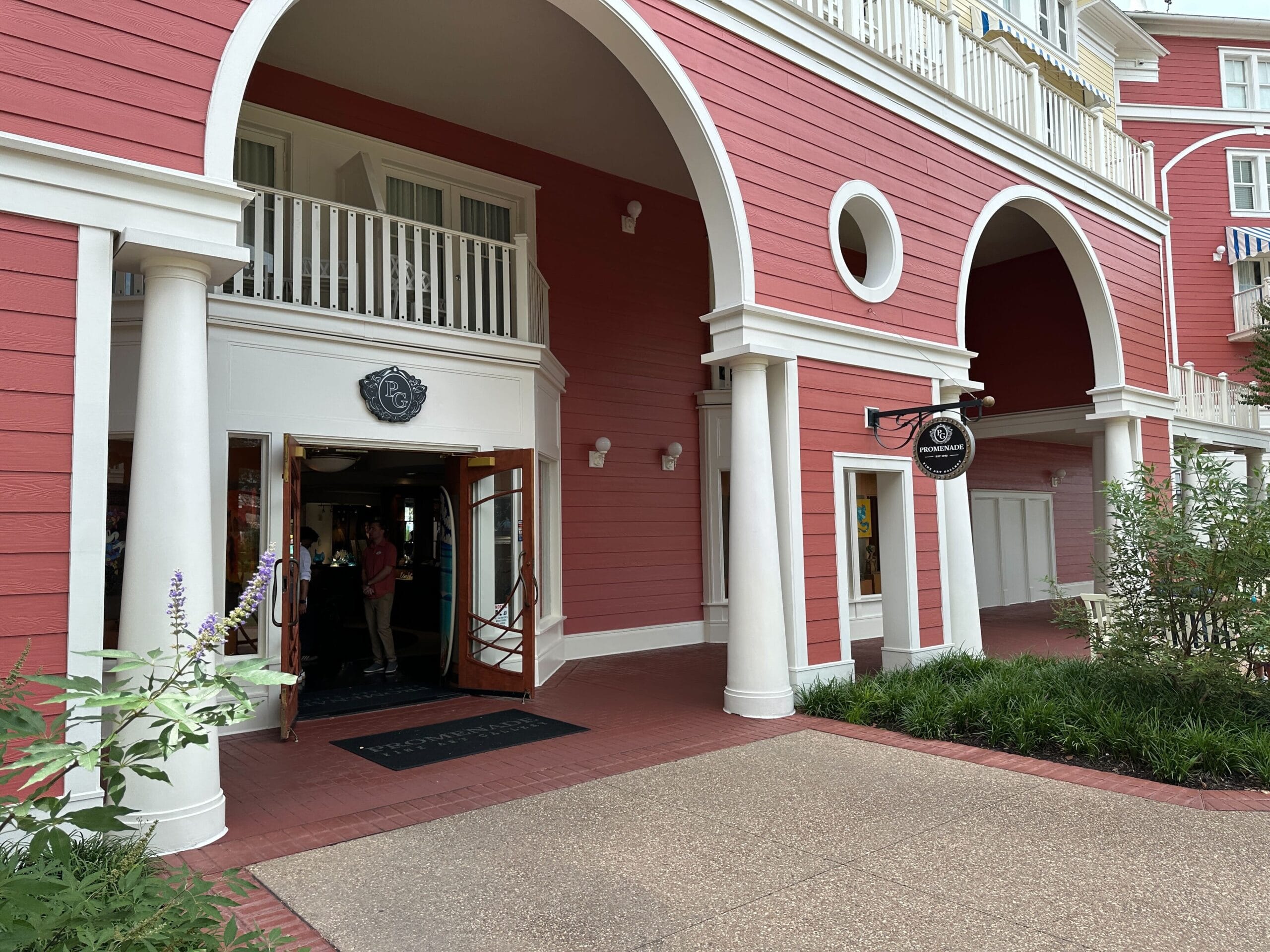 Red building with white columns and arches, open doors, greenery, and a black circular hanging sign by the entrance.