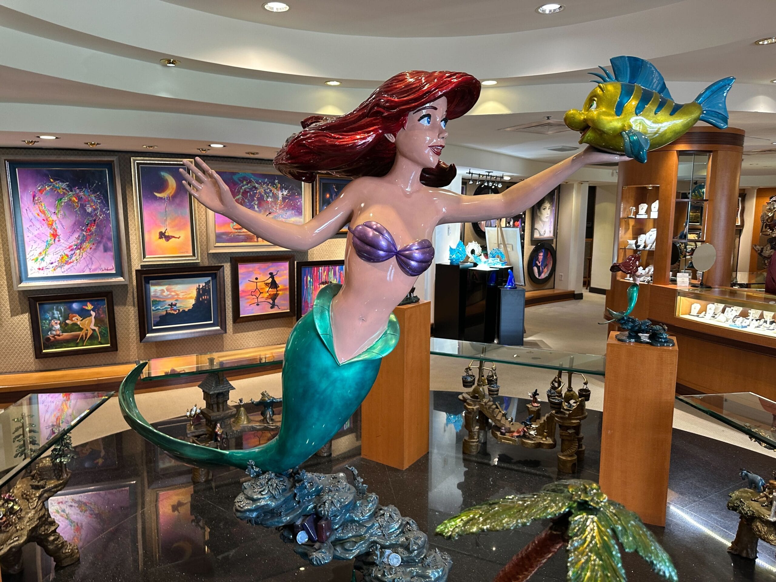 A statue of Ariel holding Flounder is displayed in an art gallery with Disney-themed paintings and jewelry.