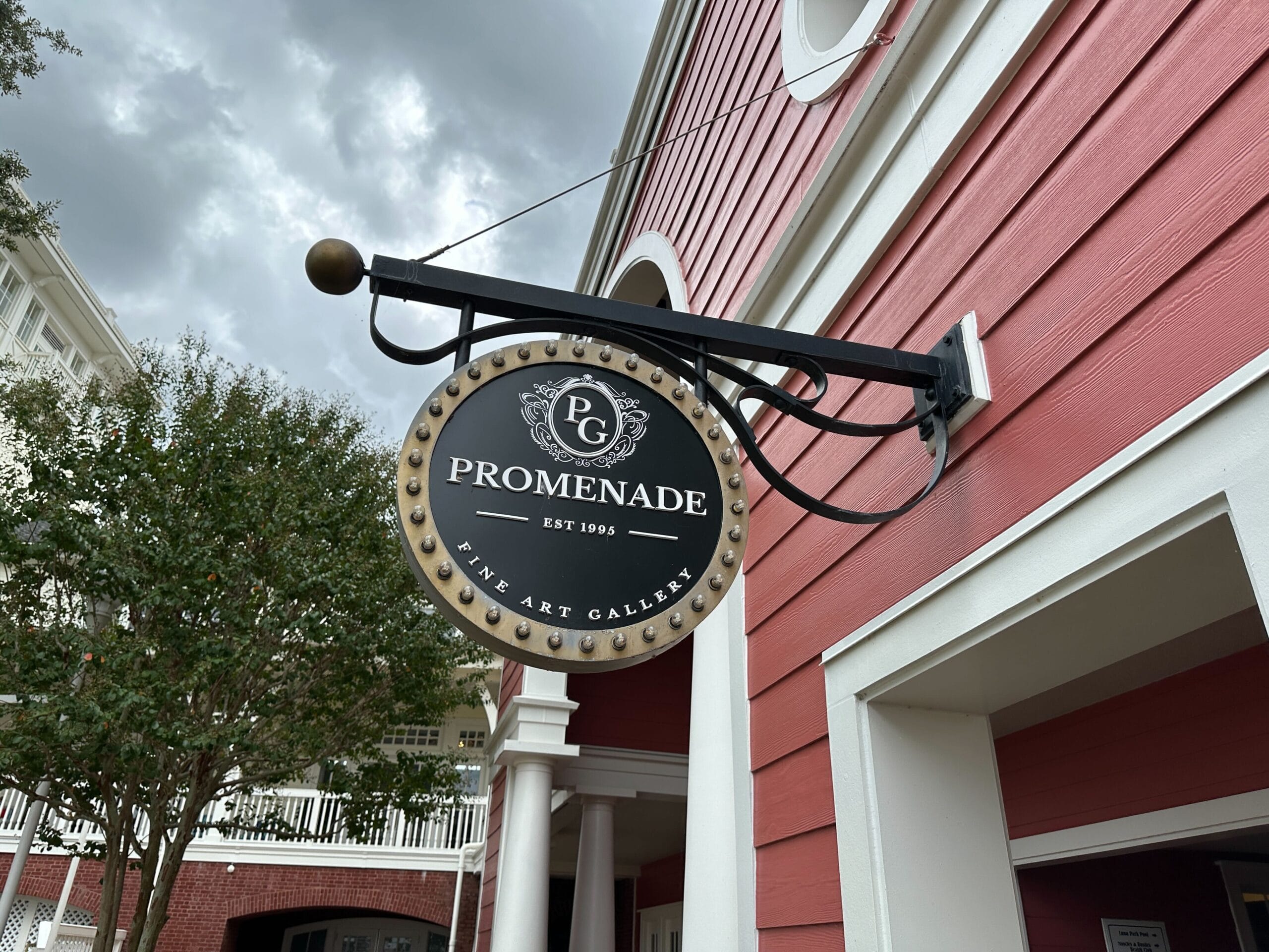 A round sign for Promenade Fine Art Gallery hangs on a red building with white trim and trees in the background.