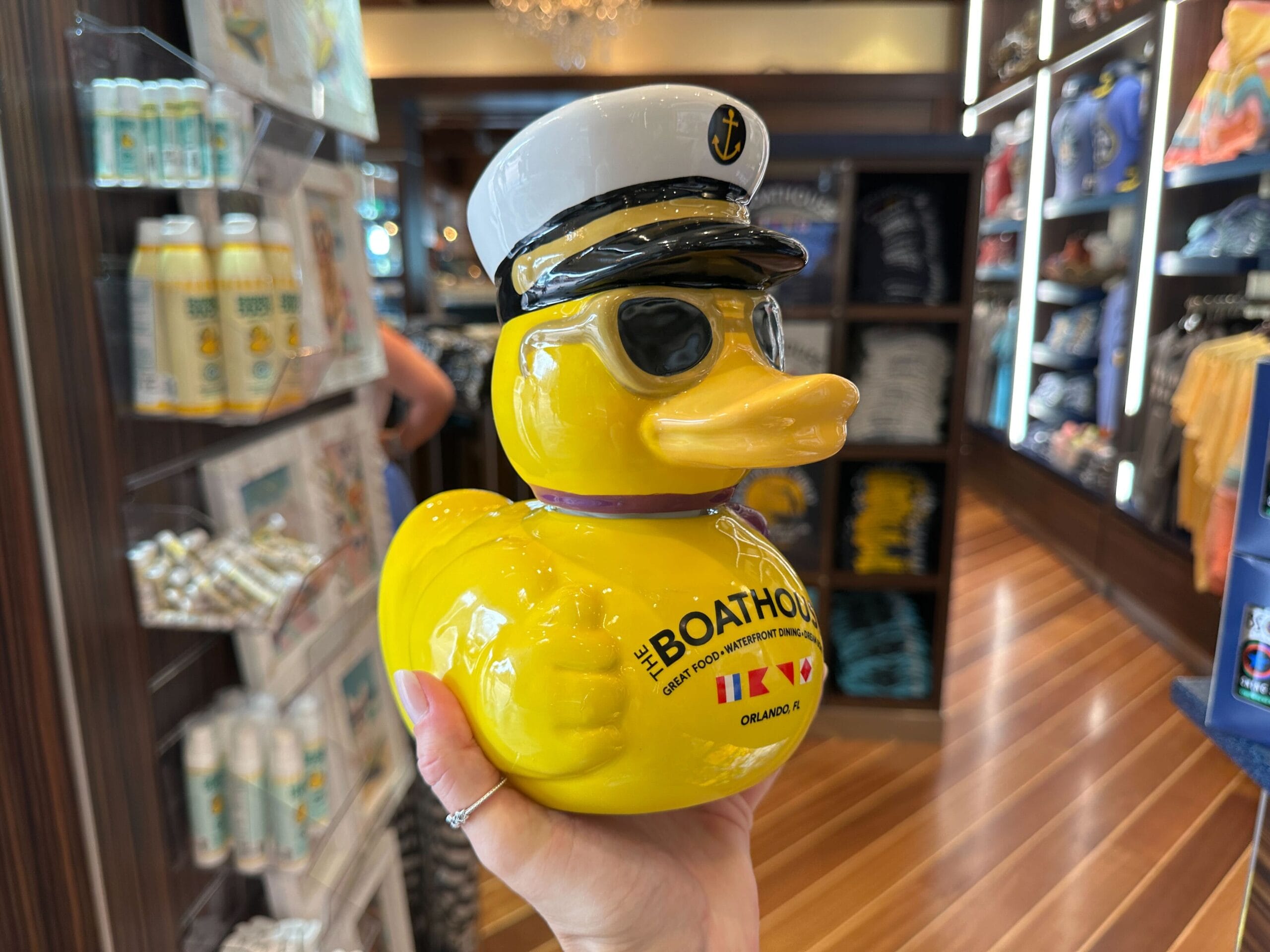 A hand holds a yellow rubber duck wearing a captain's hat and sunglasses, inside a gift shop.