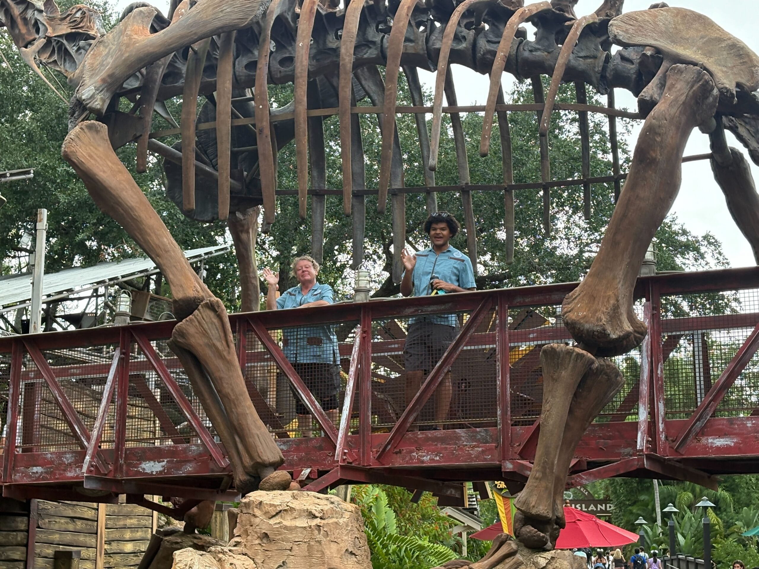 Two people wave on a bridge under a dinosaur skeleton at The Boneyard, Disney's Animal Kingdom, now closed.