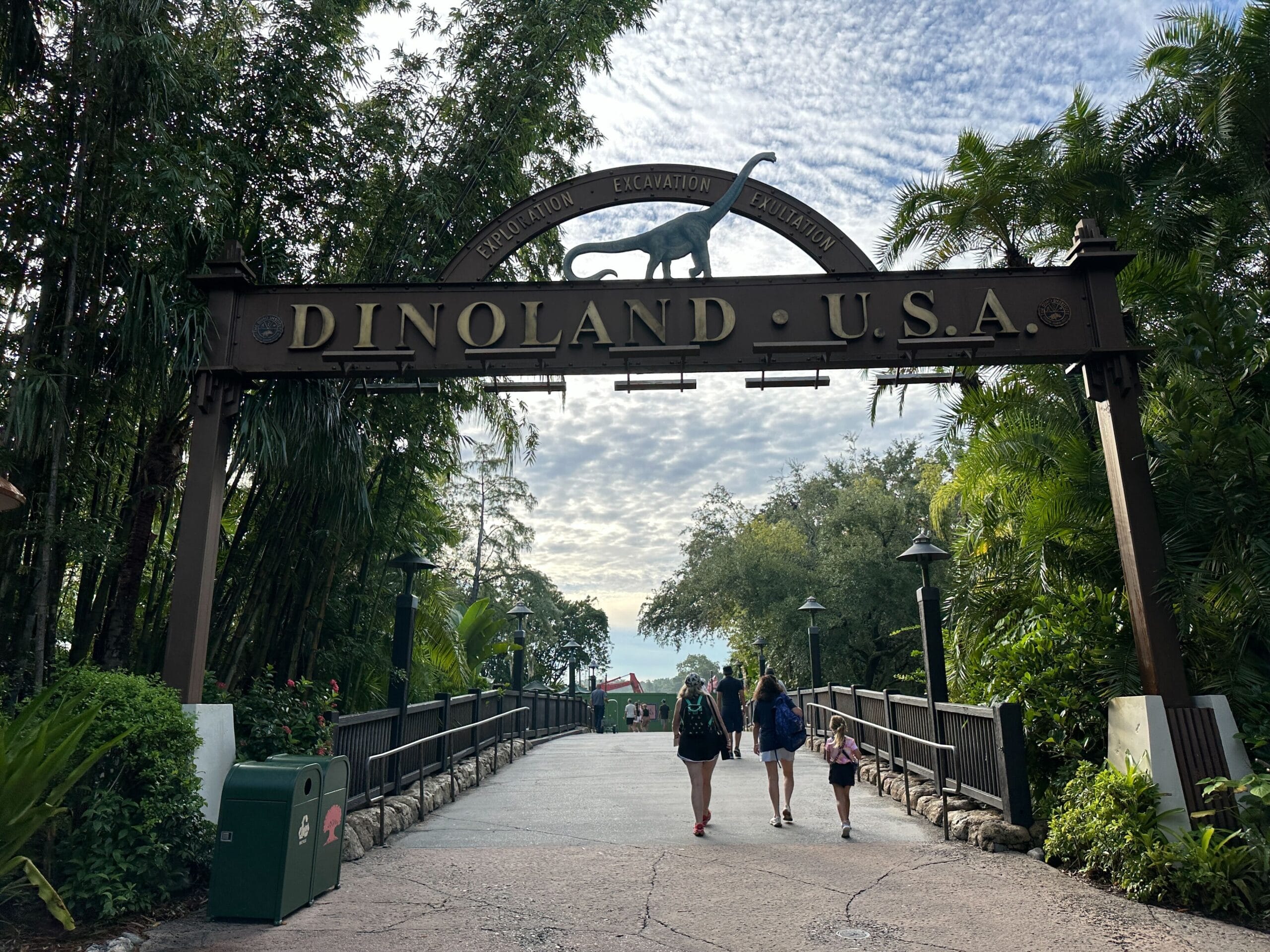 Entrance arch for Dinoland U.S.A. with a dinosaur sculpture, three people walking underneath, and lush greenery around.