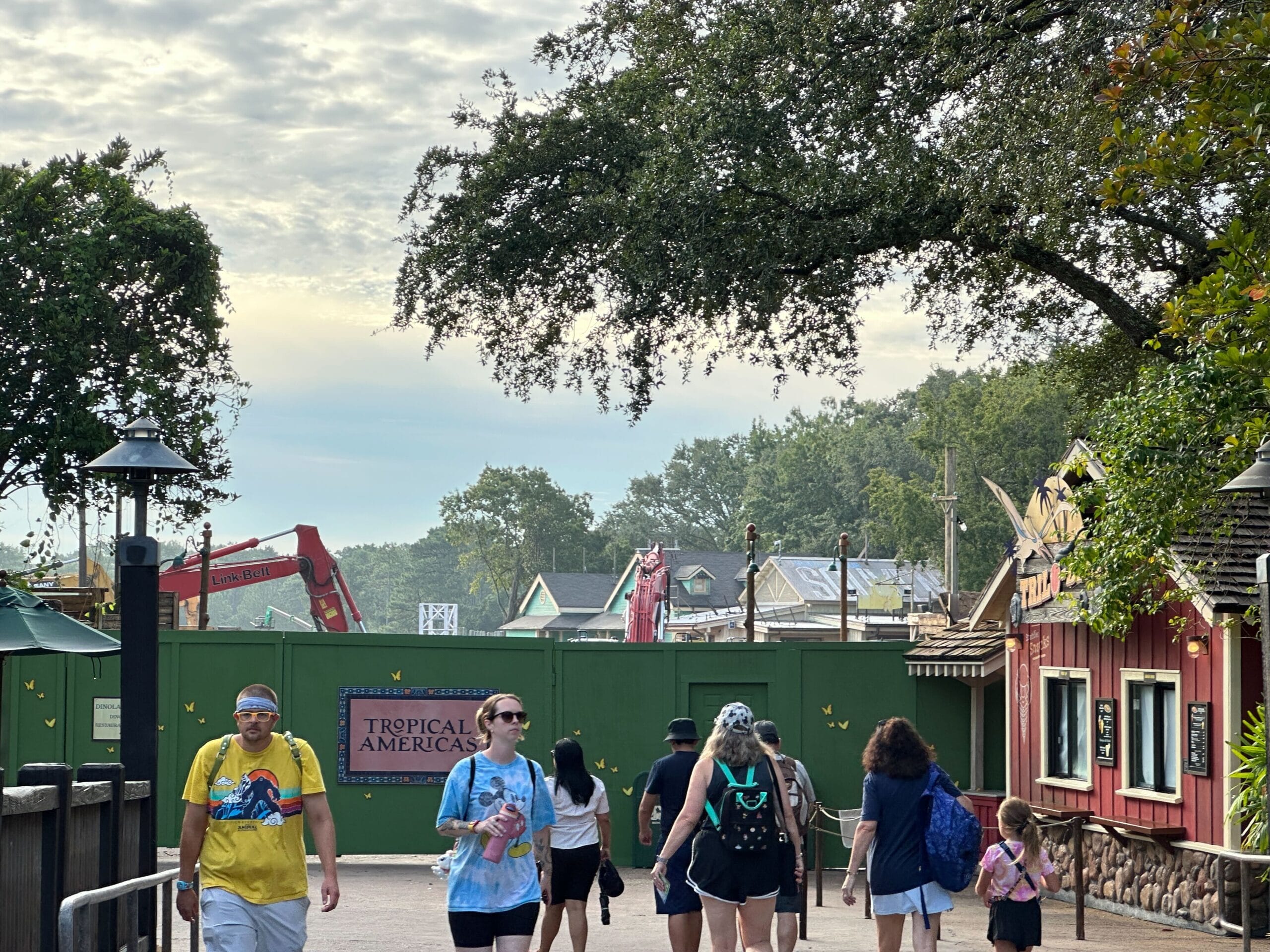 People walk past a green construction fence with a "Tropical Americas" sign at a zoo or park, under a large tree.