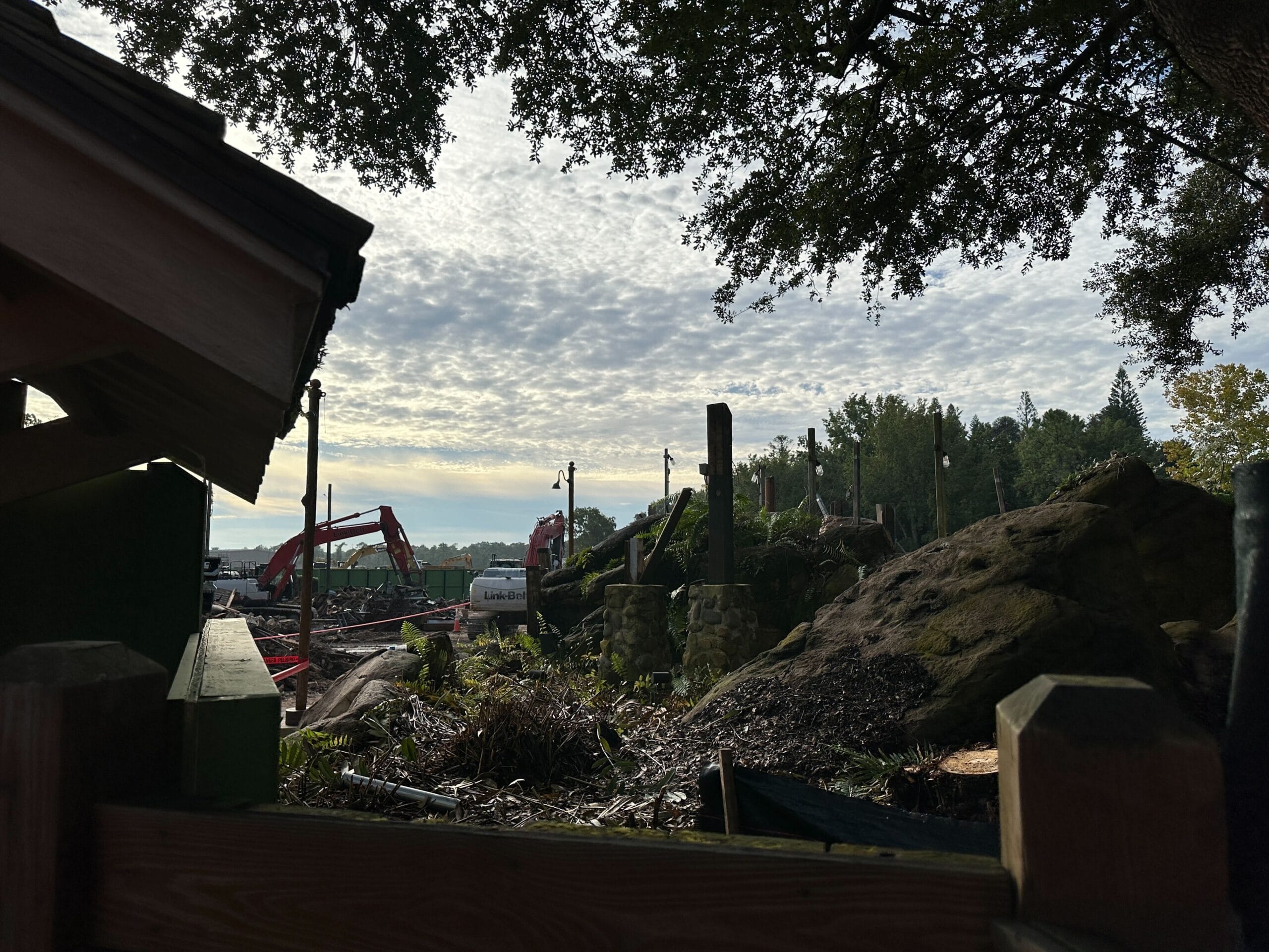 Construction vehicles and equipment at a work site surrounded by trees and rocks under a partly cloudy sky.