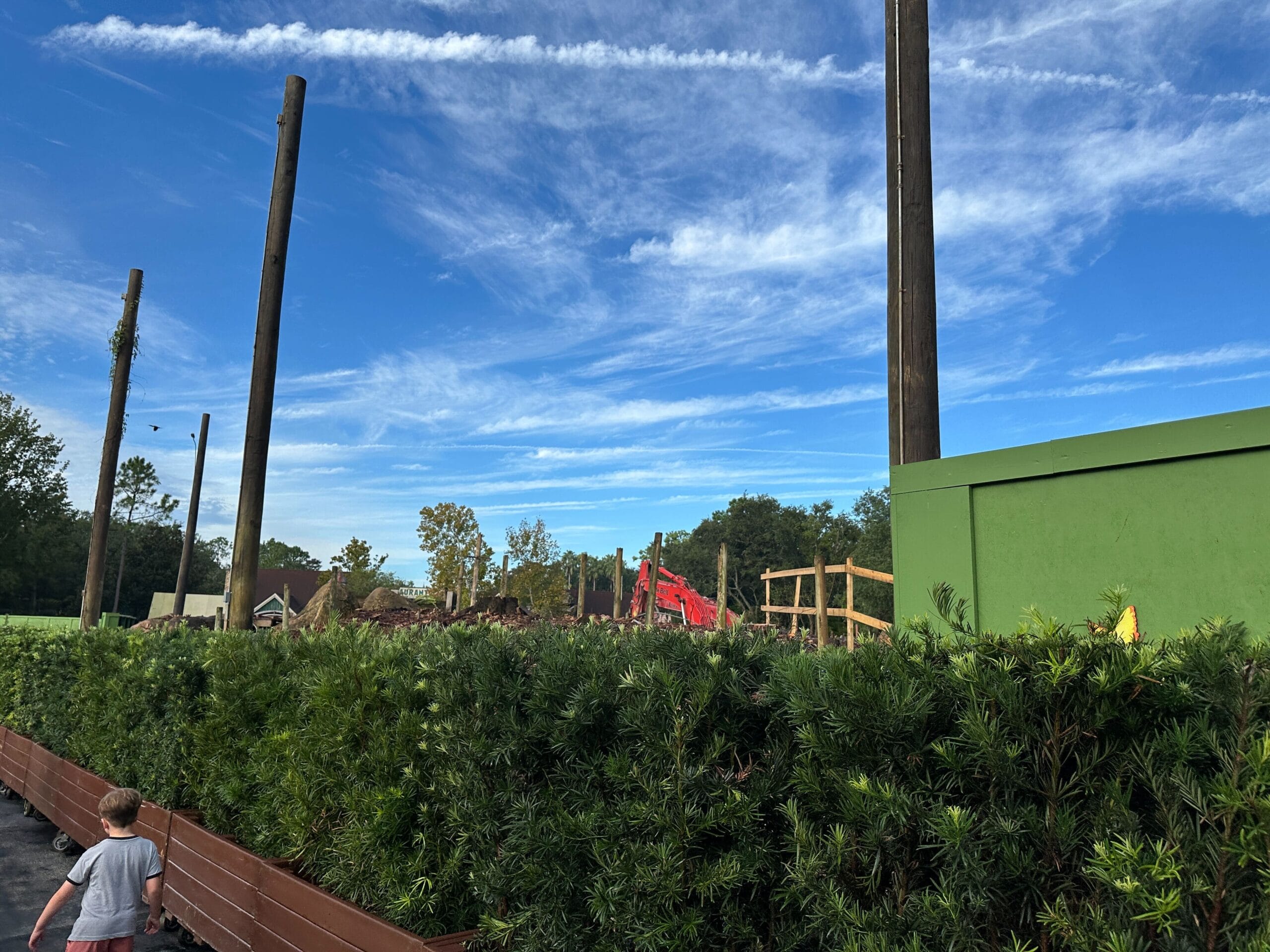 A child walks past a green fence near the removed Boneyard Bridge in Disney's Animal Kingdom under wispy blue skies.