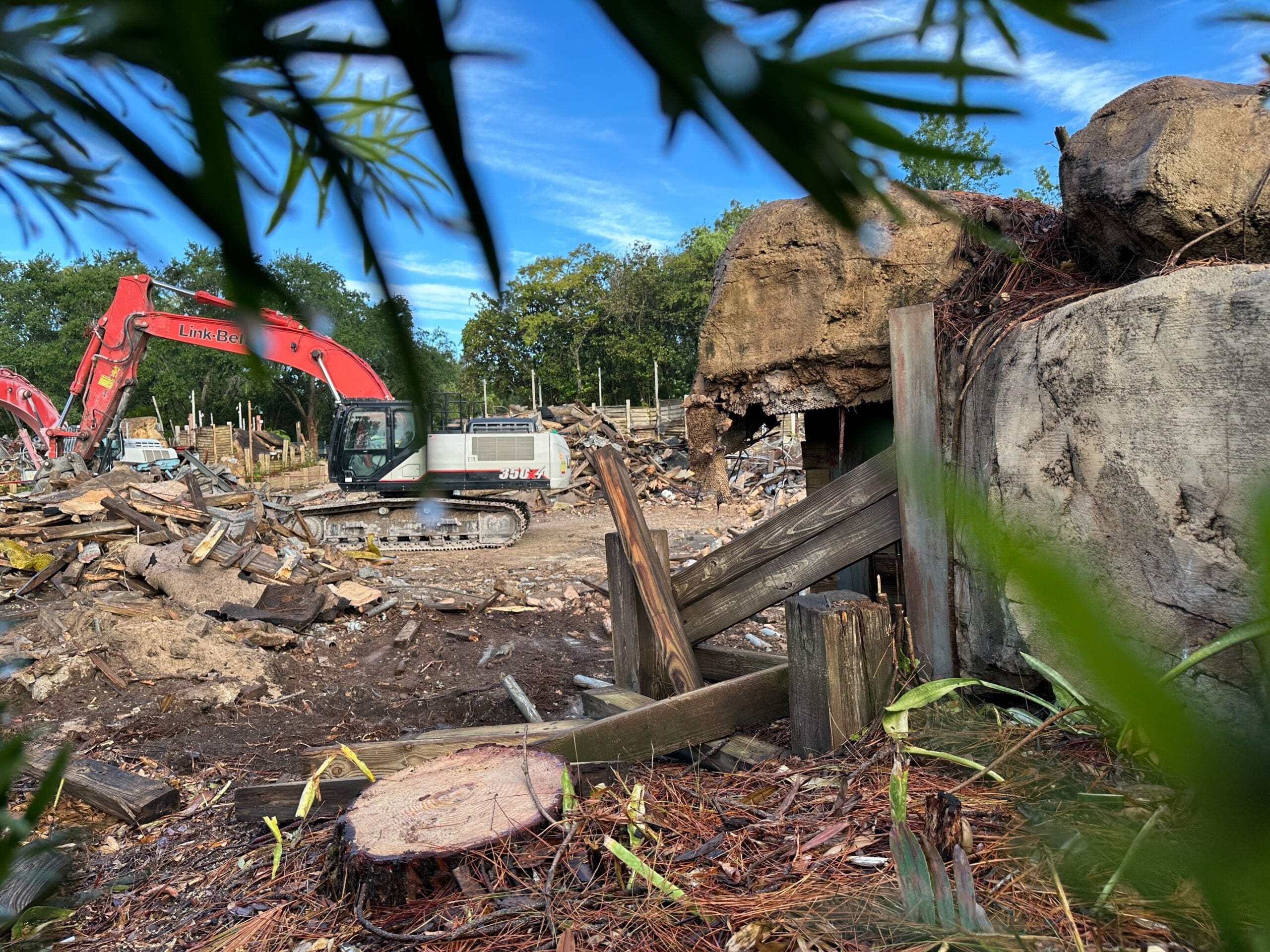 A red excavator sits where Boneyard Bridge was removed, framed by green branches and blue sky at the demolition site.