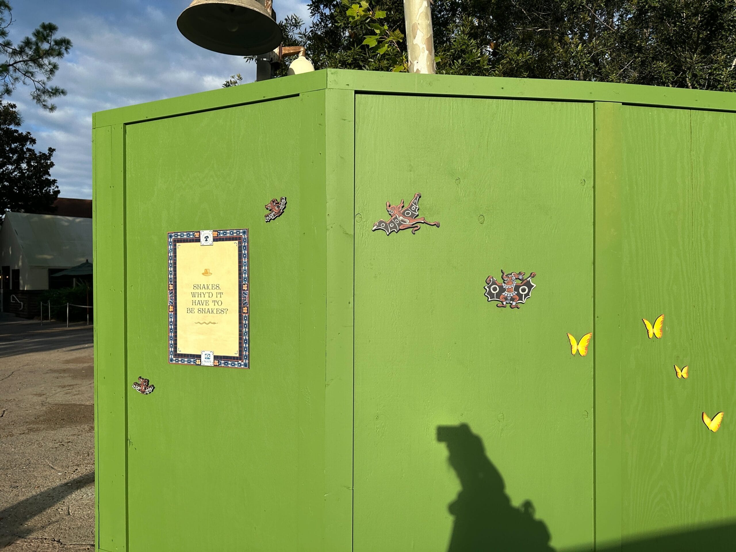 Bright green construction wall at Disney’s Animal Kingdom with cartoon bugs, butterflies, a warning sign, and a shadow in sunlight.