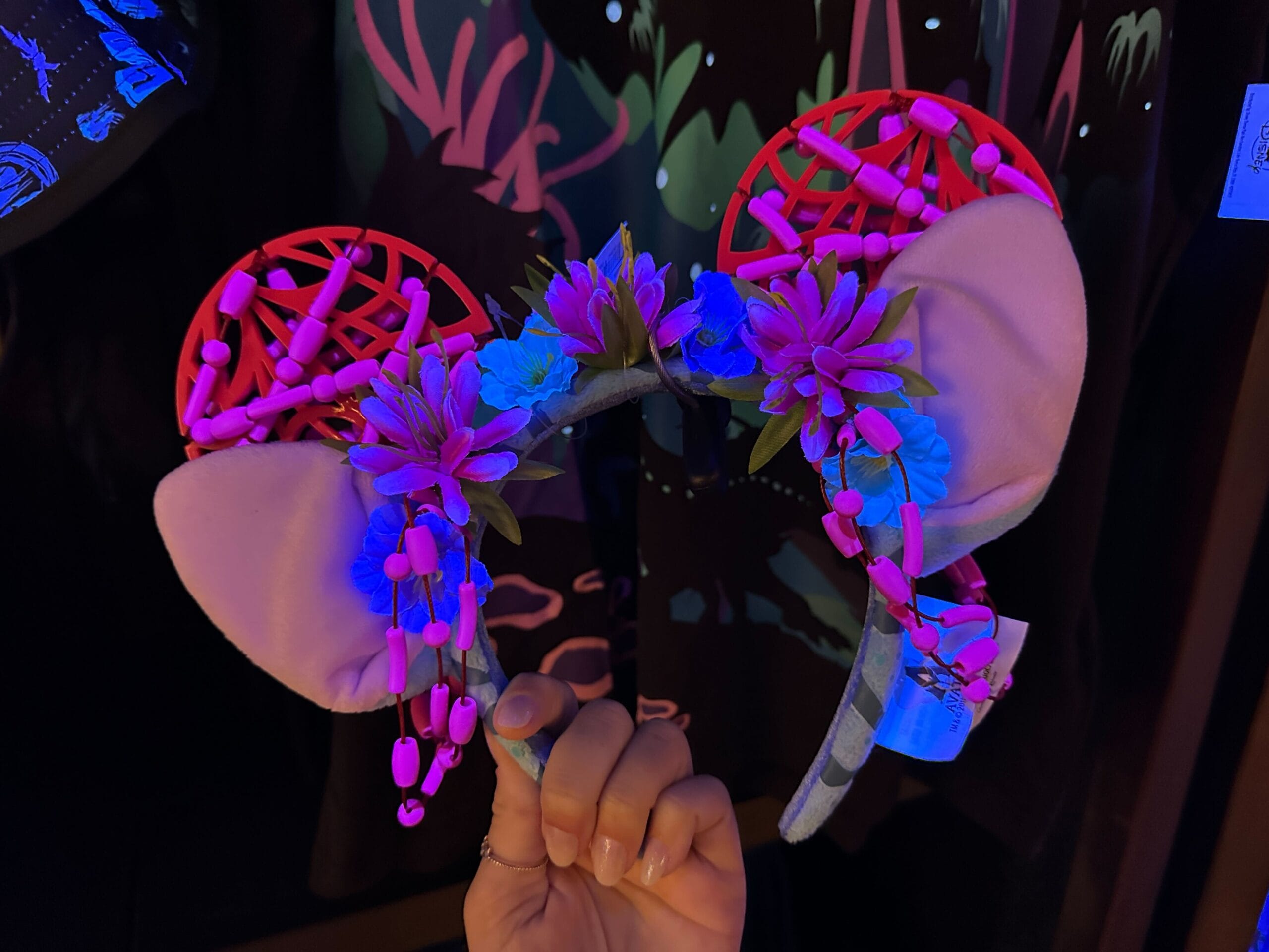 A hand holds a pink and purple glowing floral headband with animal ears, under blacklight.