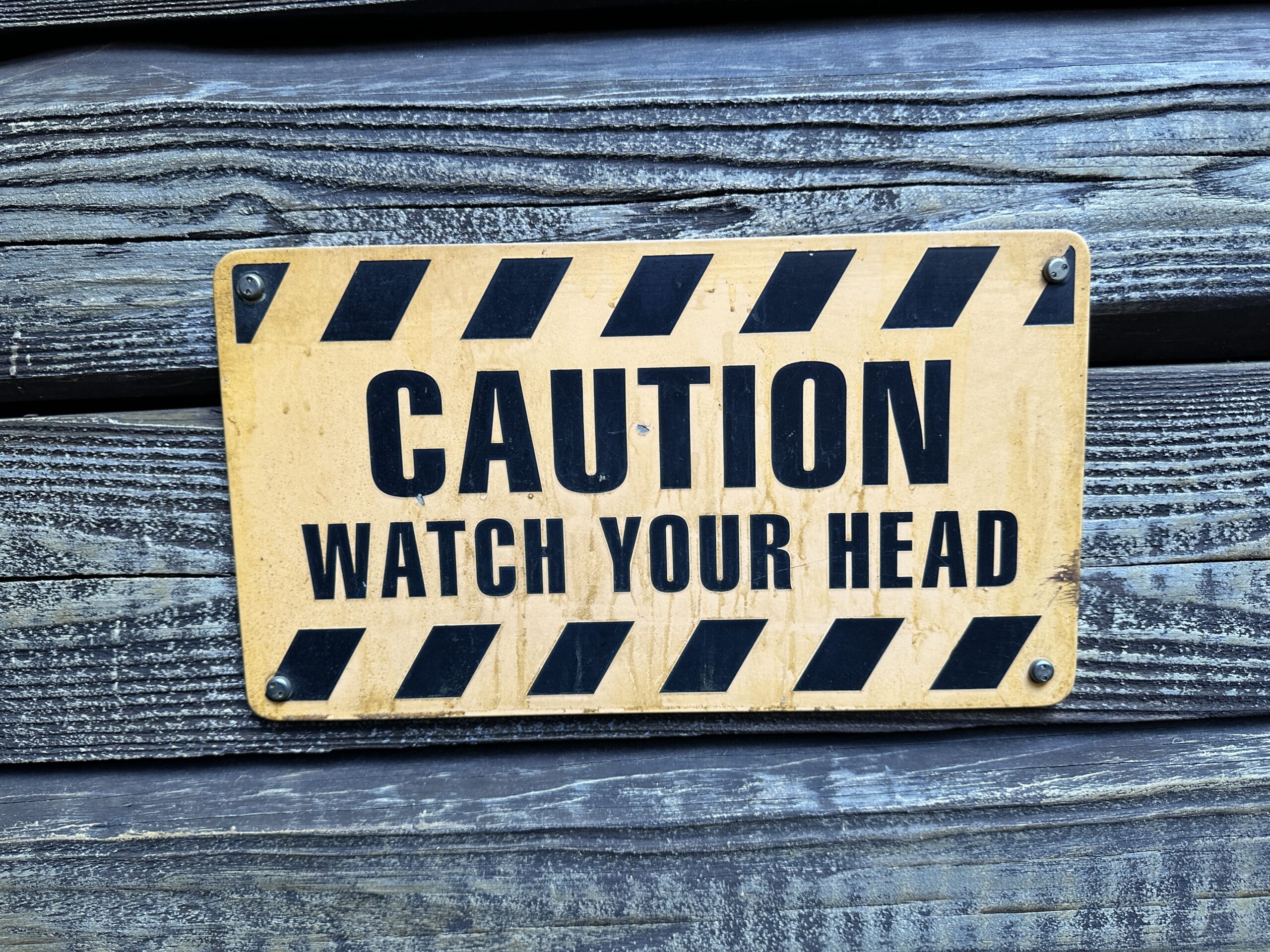 Yellow caution sign on wood wall reads "Caution Watch Your Head" with black stripes above and below the text.