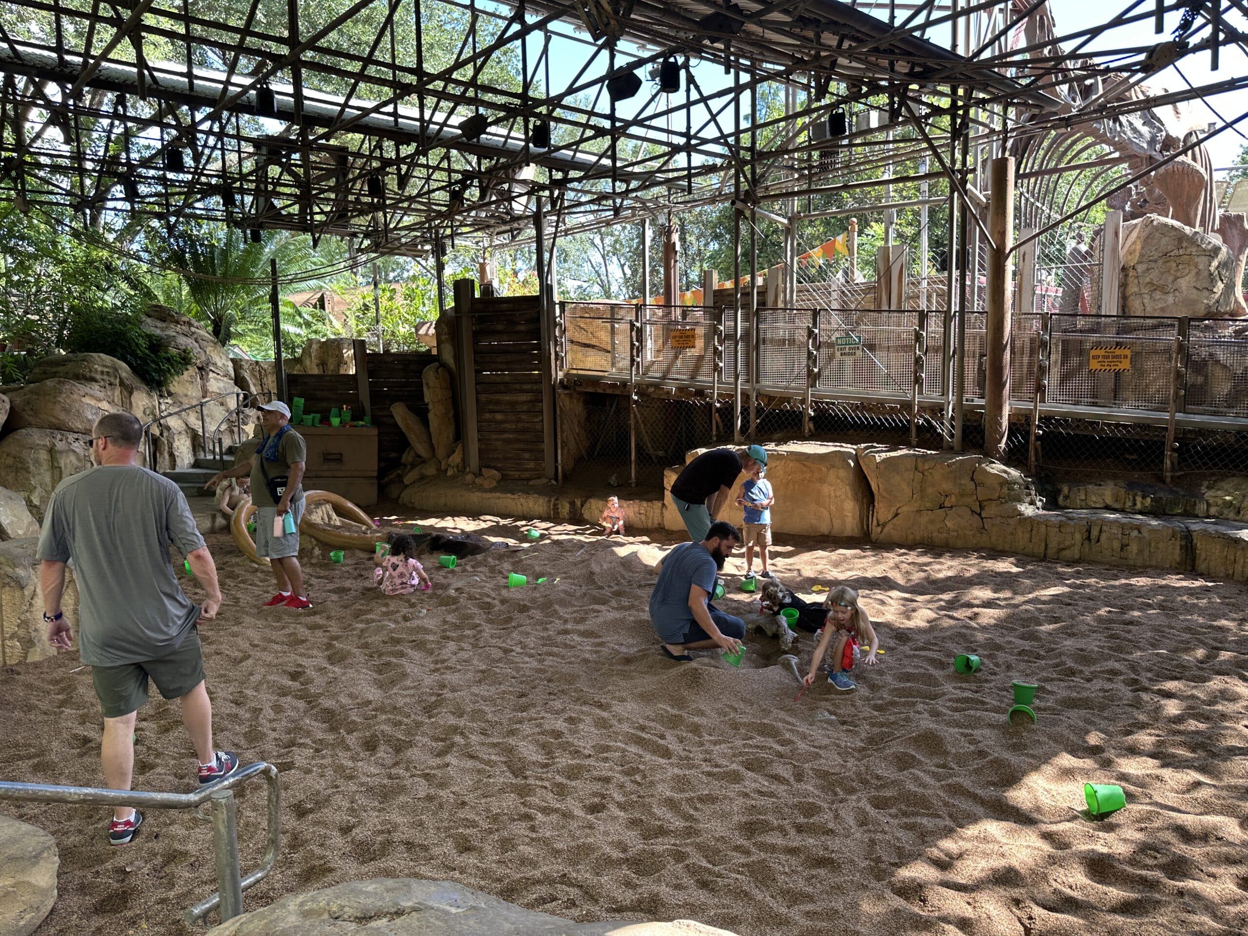 Children and adults play with green buckets in a sandy, shaded area surrounded by rocks and a metal structure.