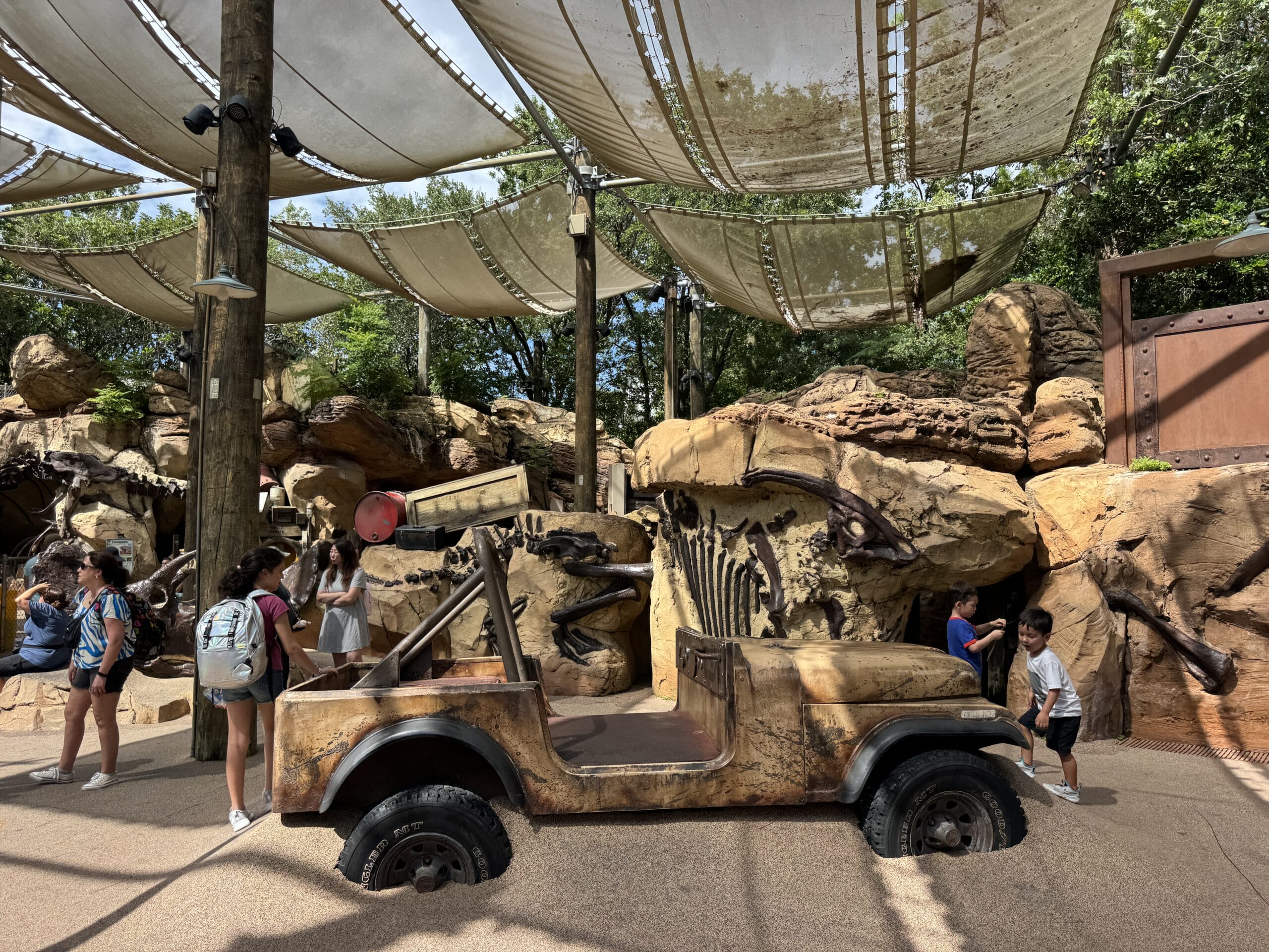 Children play around a rugged jeep and dinosaur fossils at The Boneyard in Disney's Animal Kingdom under shade canopies.