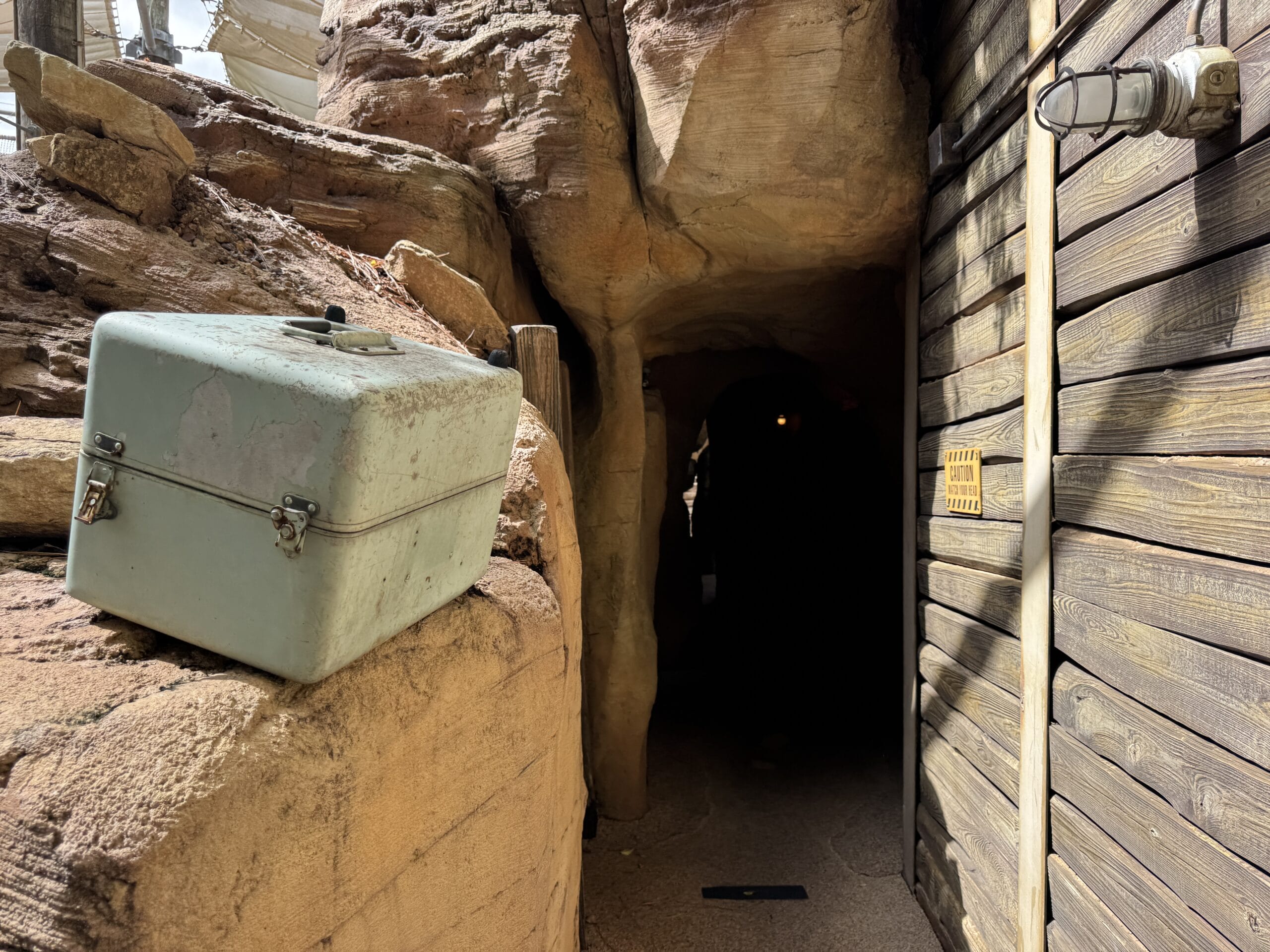 A worn metal box sits on a rock near The Boneyard, beside a dark cave entrance and vintage light fixture.