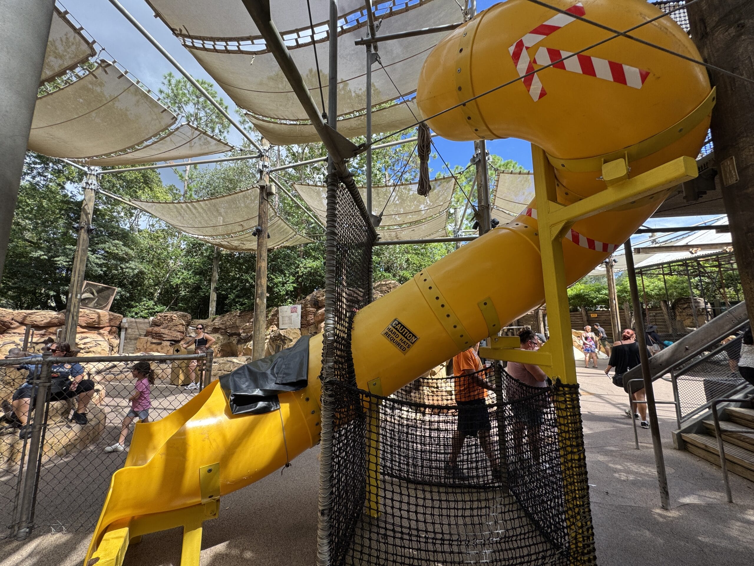 A yellow tube slide at The Boneyard in Disney's Animal Kingdom, surrounded by netting and shade sails, with people nearby.