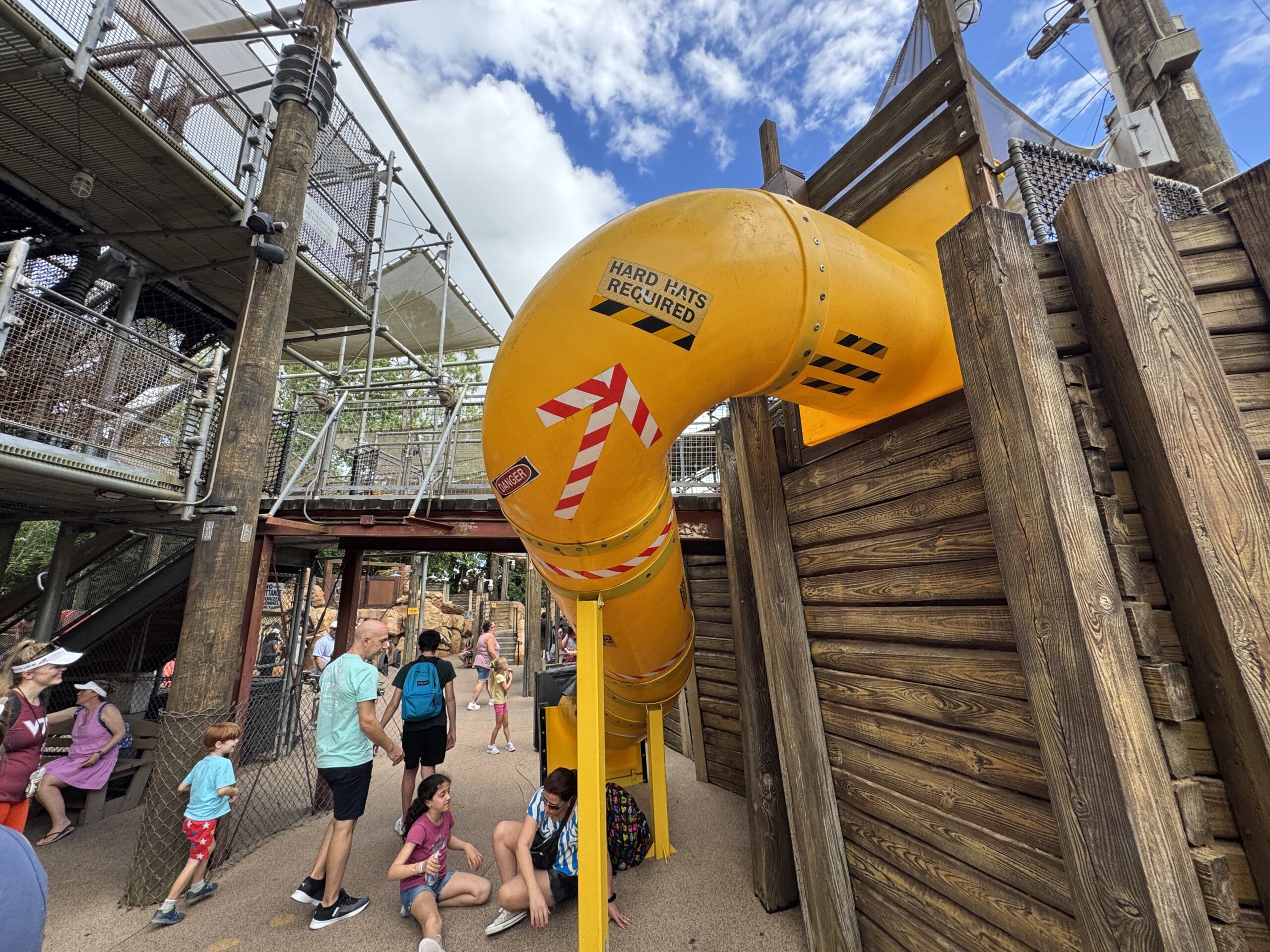 Children and adults gather near a large yellow tube slide at The Boneyard in Disney's Animal Kingdom playground.