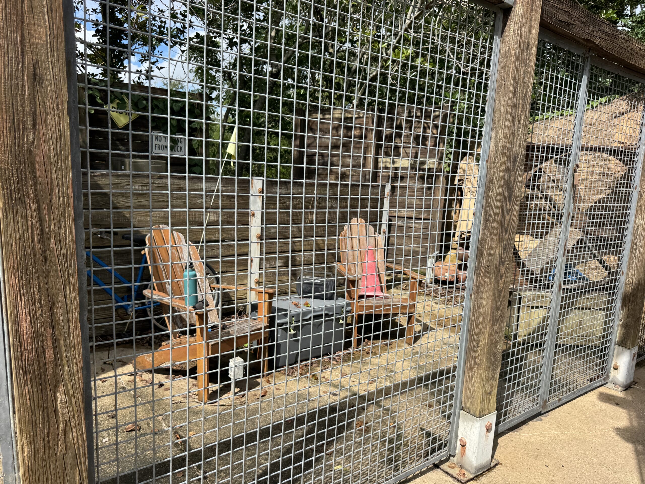 Three wooden chairs and a table sit behind a metal fence at The Boneyard, with logs and trees in the background.