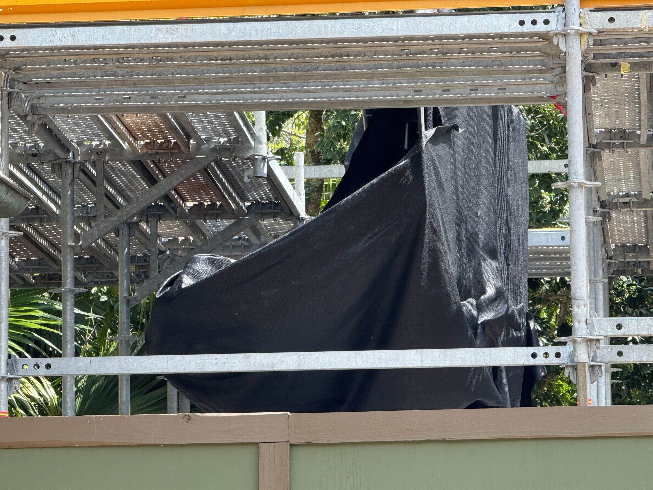 Metal scaffolding with black fabric draped over an object and trees visible in the background.