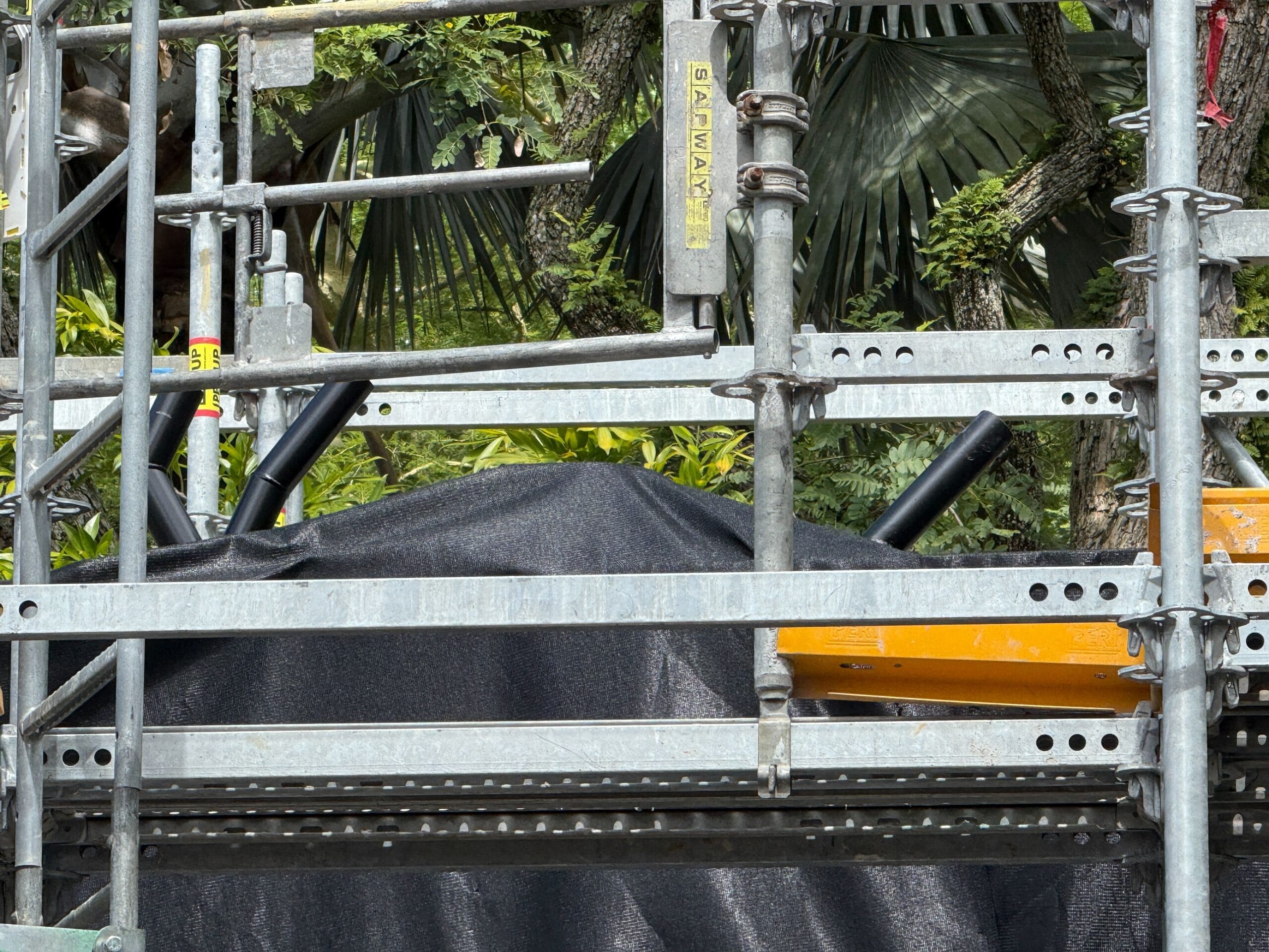 Metal scaffolding surrounds a black tarp and orange structure, with trees and greenery visible in the background.