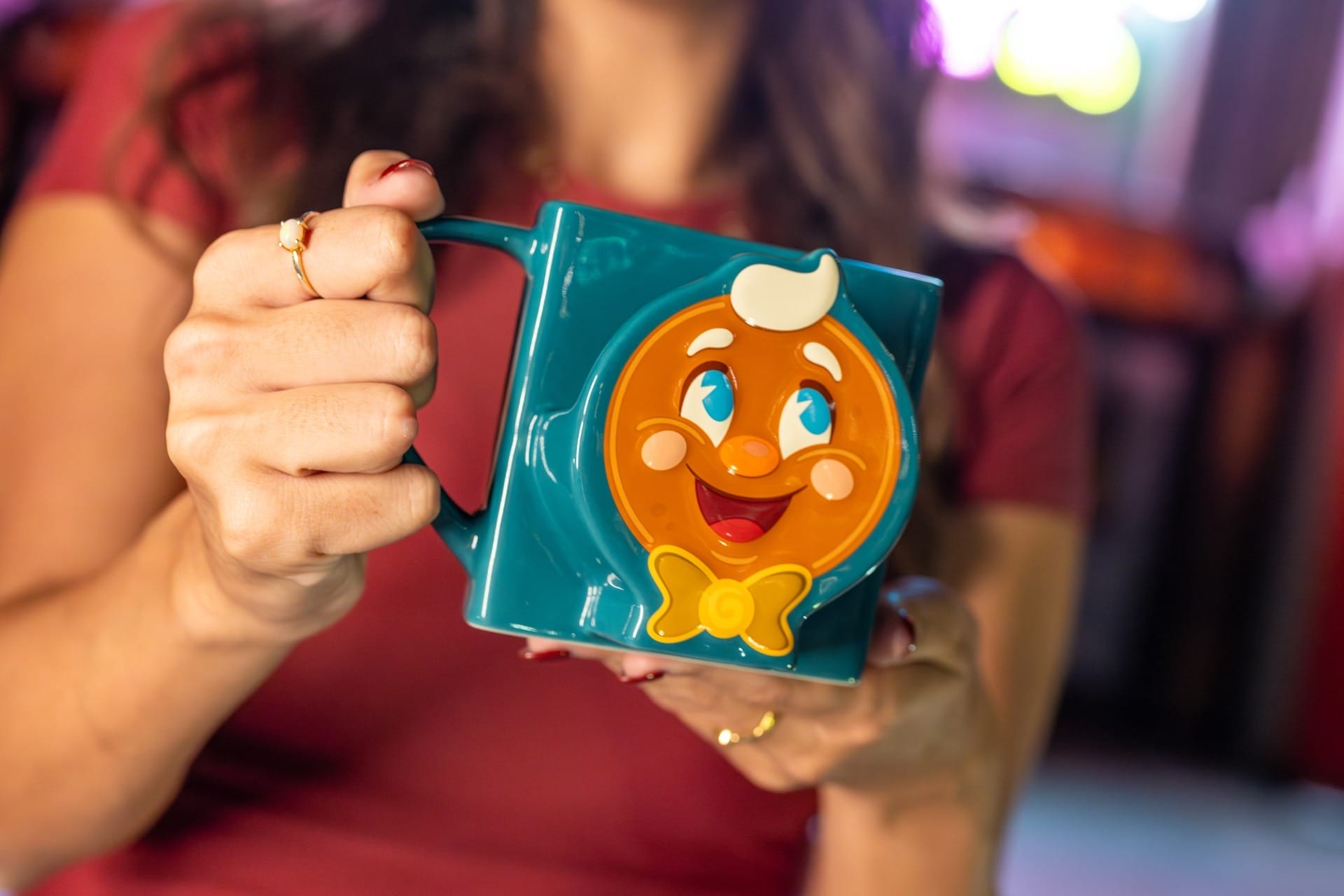 A person holds a teal mug with Ollie the Gingerbread Man, wearing a red shirt—perfect 2025 merchandise.