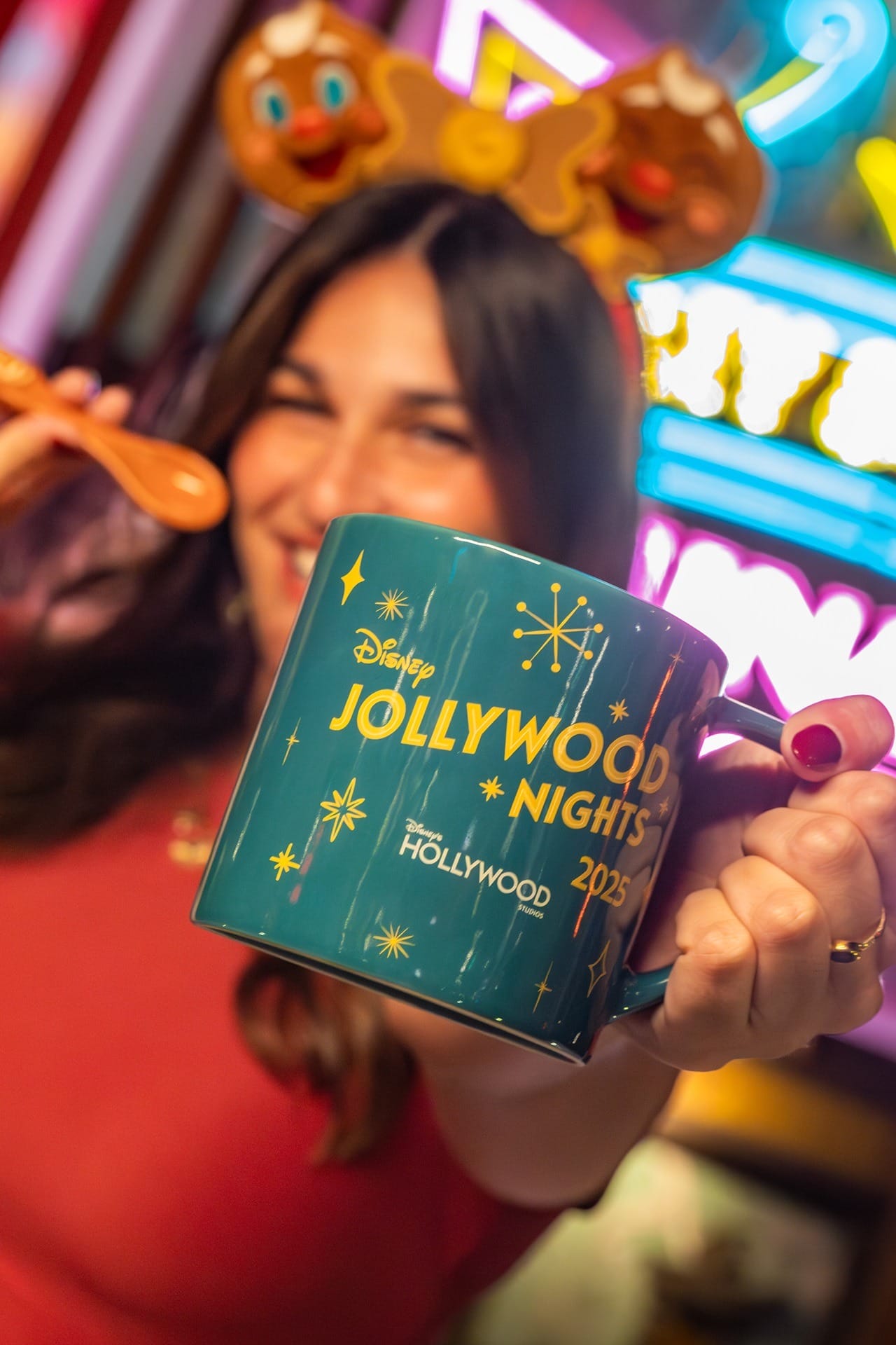 Woman holding Disney Jollywood Nights 2025 mug, wearing Ollie the Gingerbread Man headband, colorful lights in background.