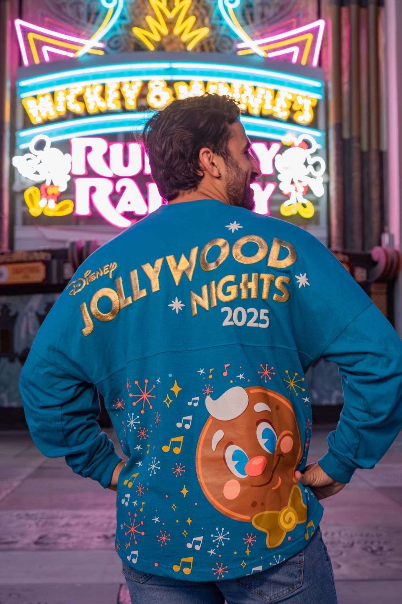 A man in Disney Jollywood Nights 2025 merchandise with Ollie the Gingerbread Man stands before glowing neon signs.
