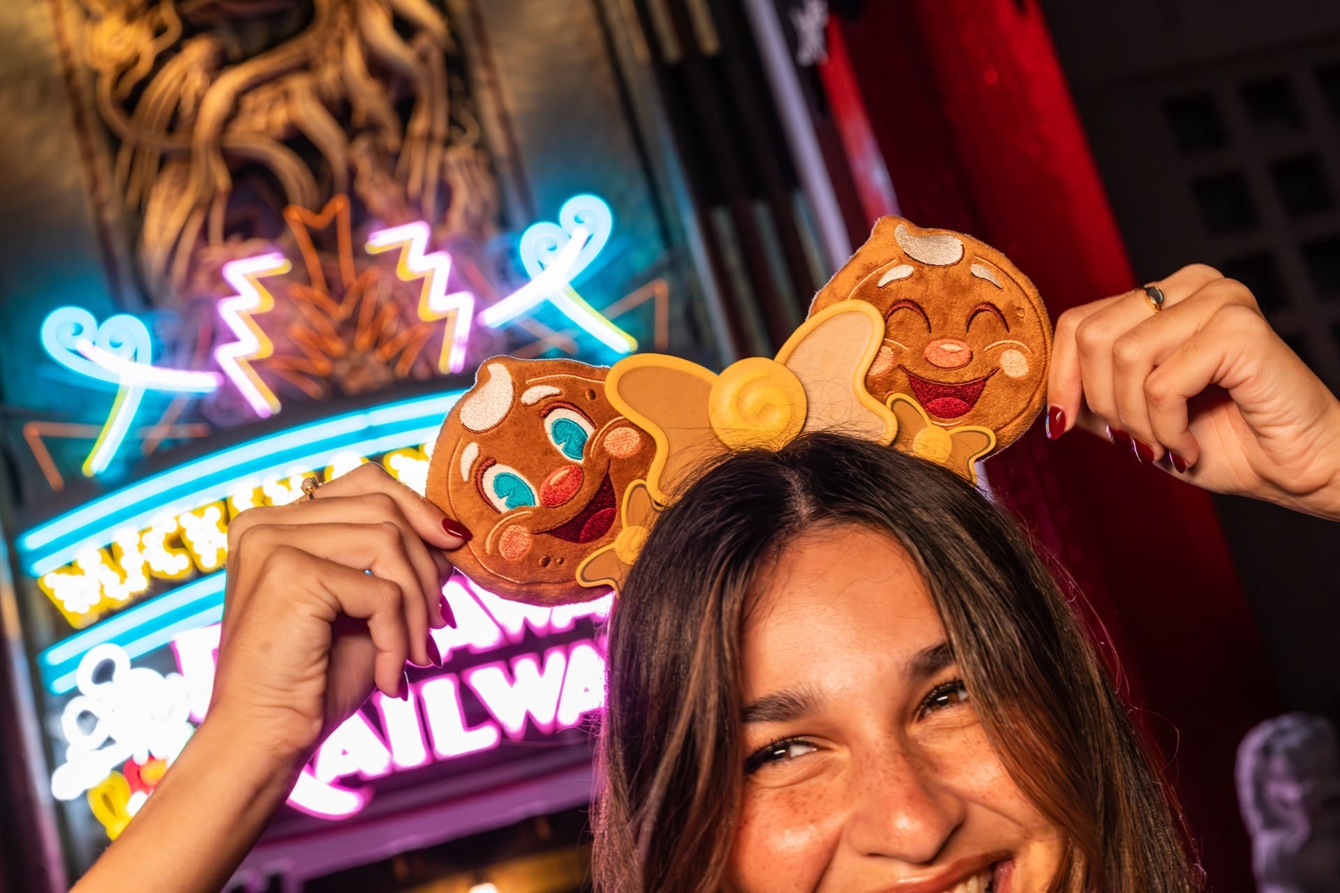 Smiling woman wearing Disney Jollywood Nights gingerbread Mickey ears in front of a bright neon sign at night.