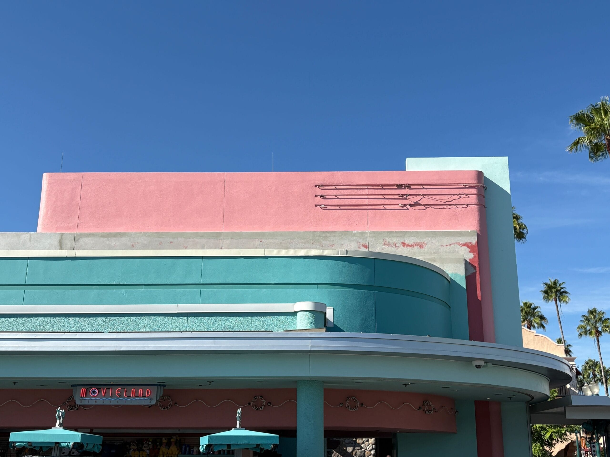 A pink and teal building with a faded sign under a clear blue sky and palm trees in the background.
