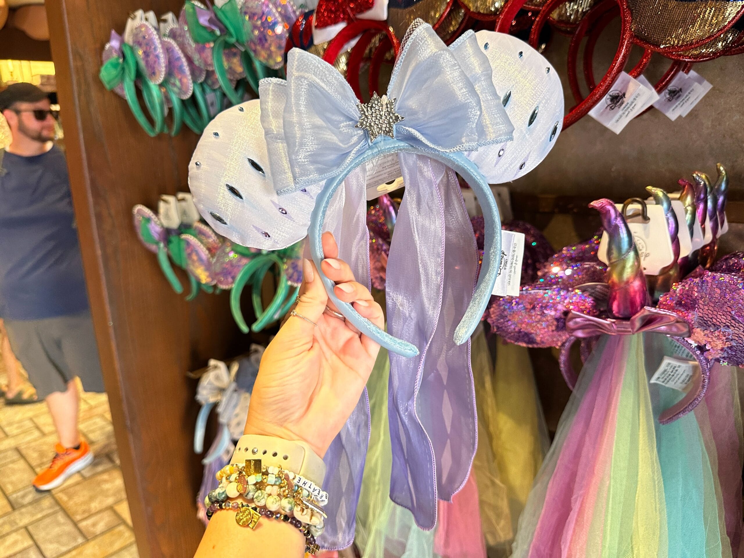 A hand holds a sparkly blue and purple Frozen 2 Elsa Ear Headband with a big bow and veil in a store display of headbands.