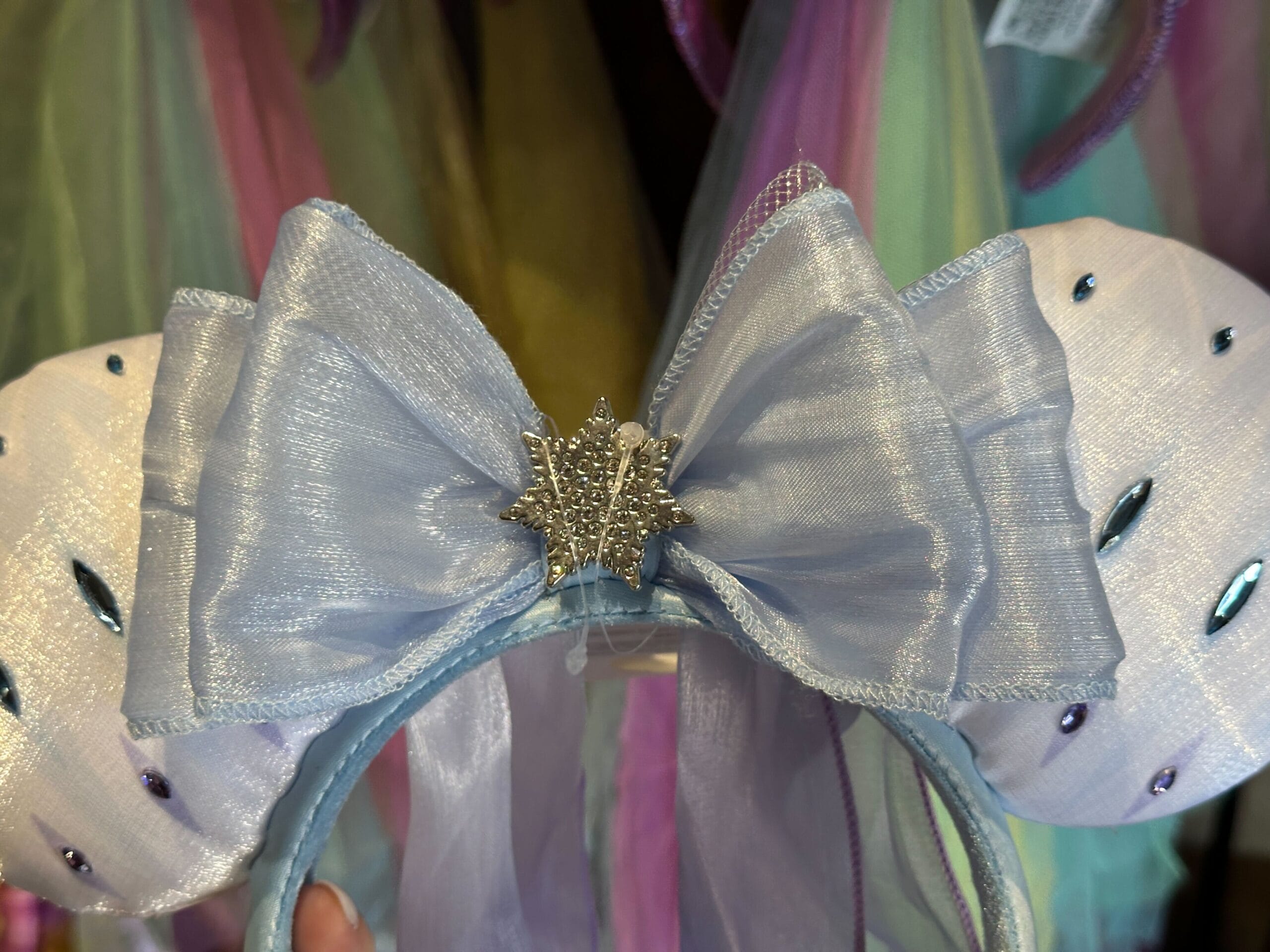 Light blue Elsa Ear Headband with a shimmery bow and jeweled snowflake, inspired by Frozen 2, pastel colors in the background.