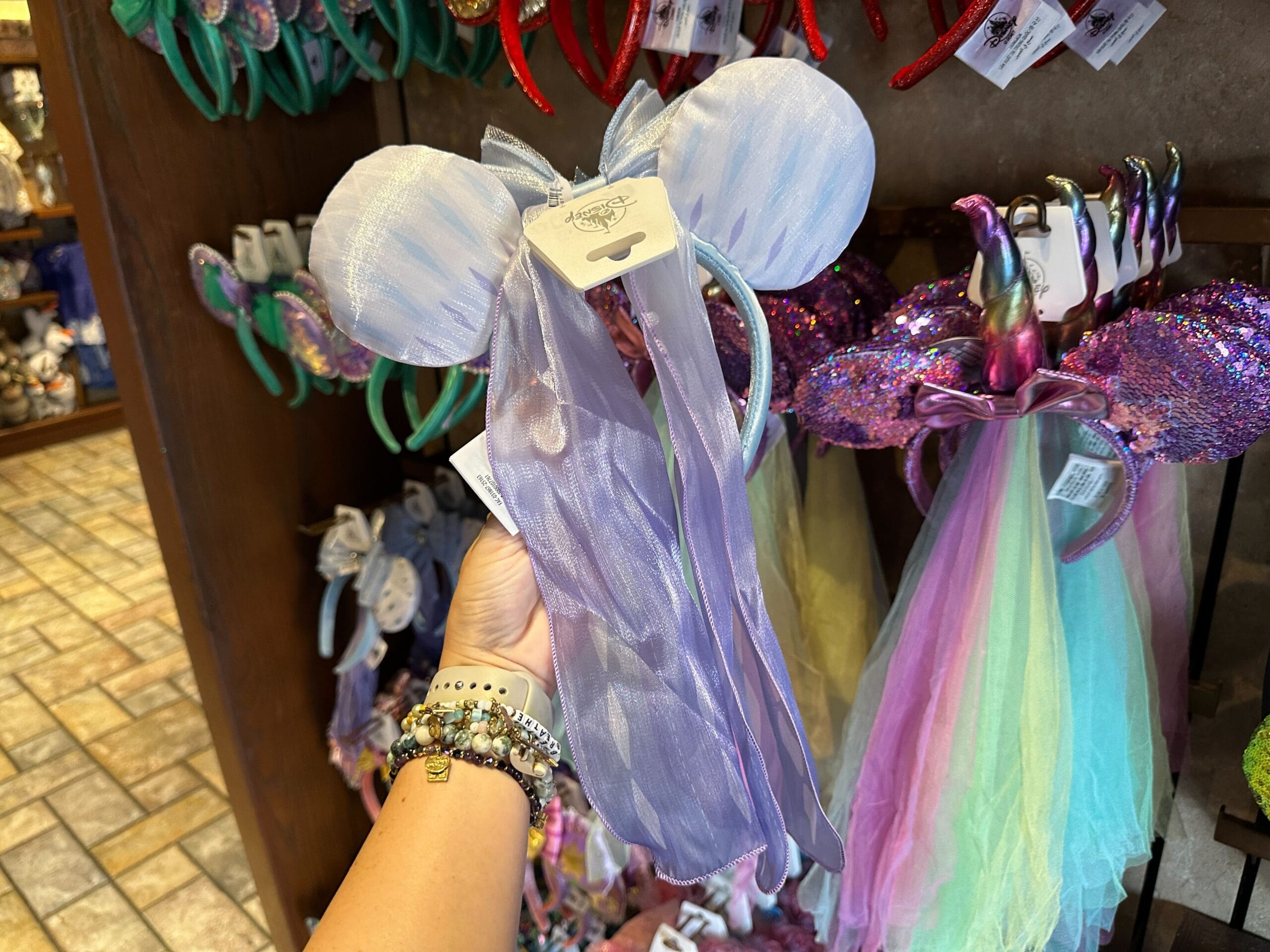 A hand holds a pastel purple Frozen 2 Elsa Ear Headband in a store at Walt Disney World, surrounded by colorful accessories.