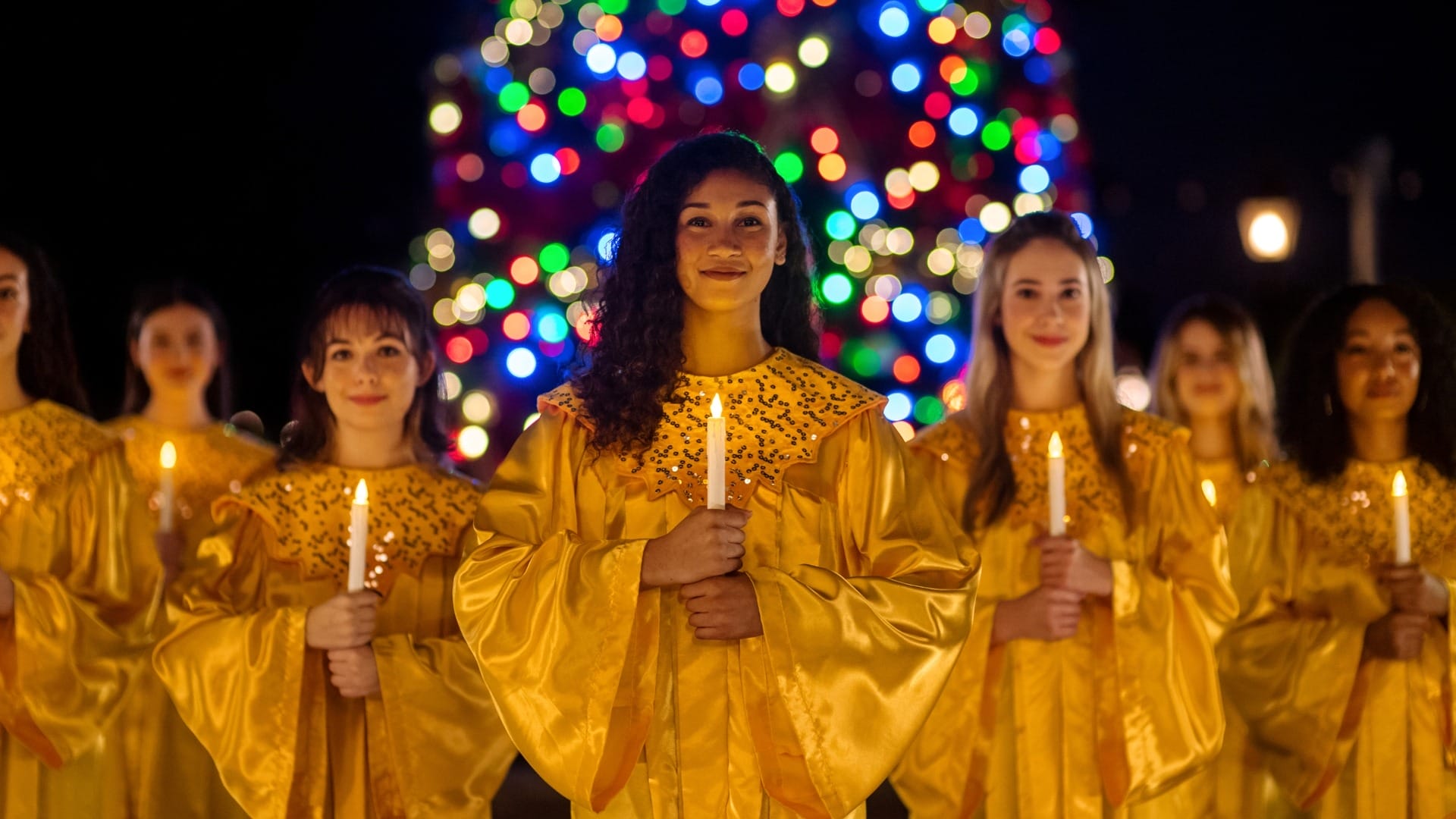 A group of women in gold robes hold candles during the Candlelight Processional at EPCOT, near a glowing Christmas tree.