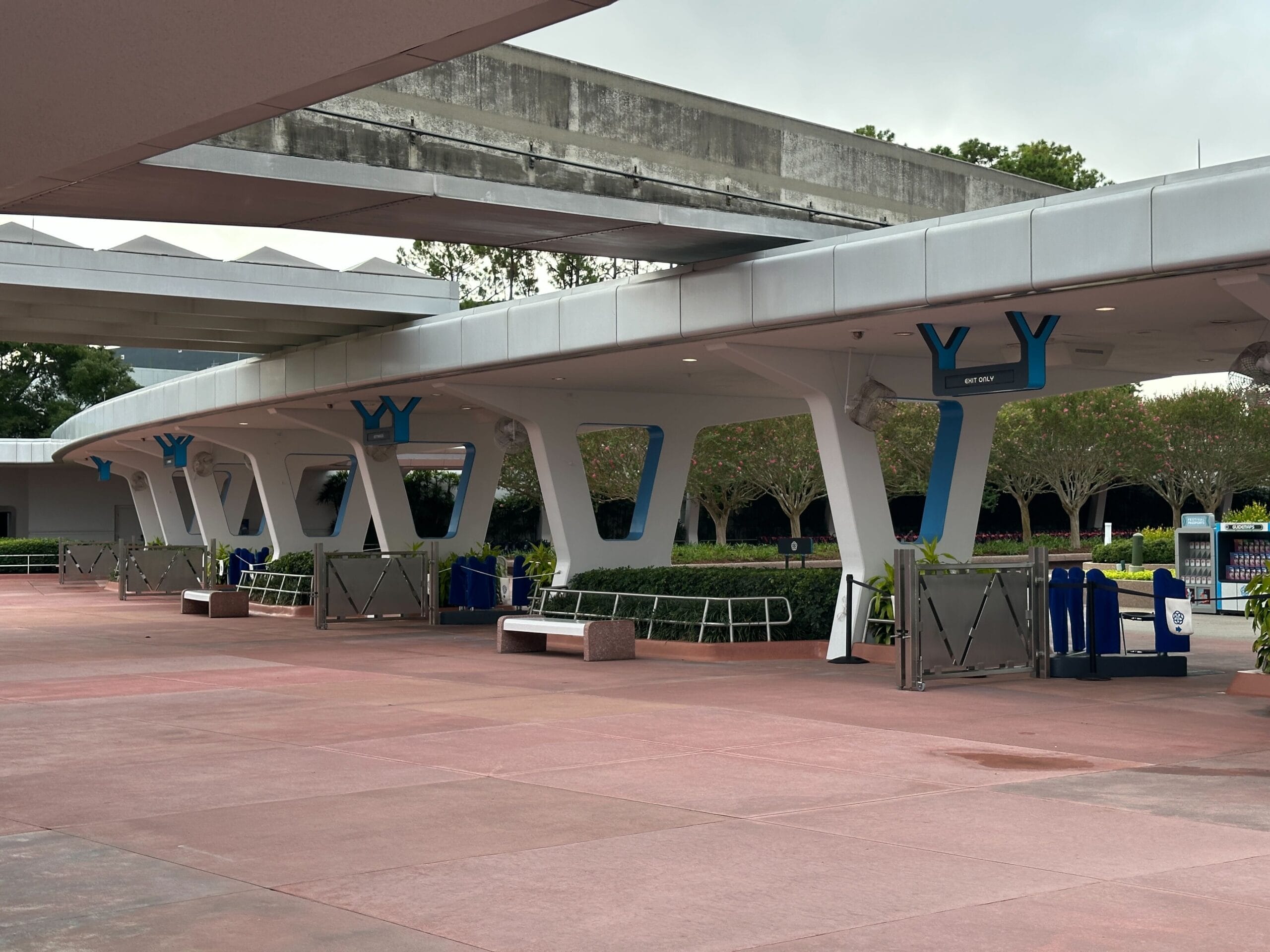 A nearly empty walkway with modern white pillars and blue signs, featuring some benches and greenery in the background.